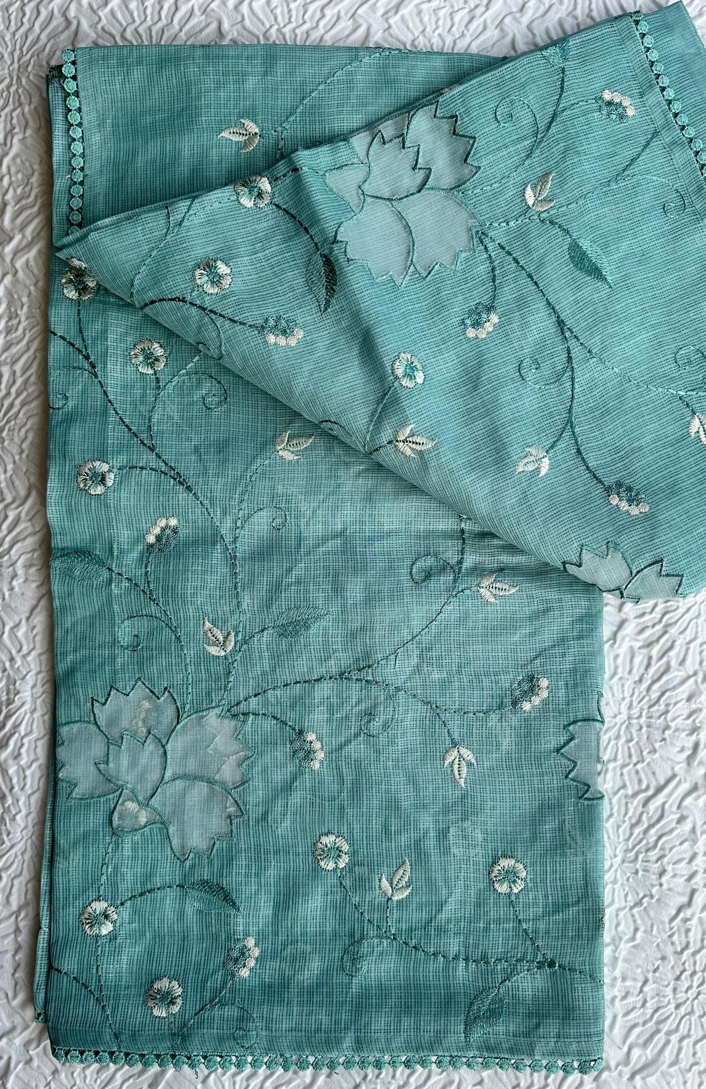 Beautiful sage green kota cotton saree with delicate white and green applique floral work scattered across the fabric, featuring decorative border trim, displayed on white textured background