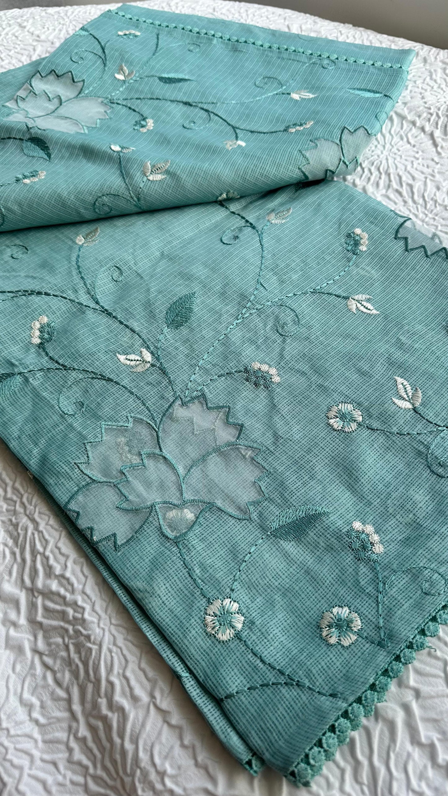 Beautiful sage green kota cotton saree with delicate white and green applique floral work scattered across the fabric, featuring decorative border trim, displayed on white textured background