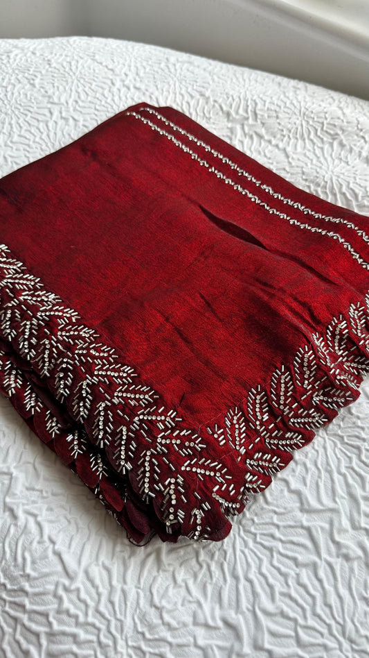 Luxurious deep red silk saree with intricate silver thread embroidery and beadwork featuring elaborate leaf patterns along the border, displayed on white textured fabric