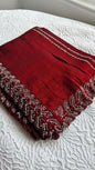 Luxurious deep red silk saree with intricate silver thread embroidery and beadwork featuring elaborate leaf patterns along the border, displayed on white textured fabric