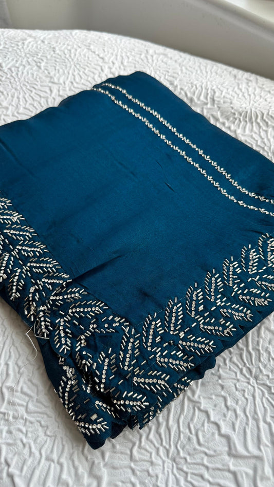 Elegant teal blue barberry saree with intricate silver gota work embroidery featuring detailed leaf patterns along the border, beautifully displayed on white textured background