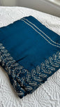 Elegant teal blue barberry saree with intricate silver gota work embroidery featuring detailed leaf patterns along the border, beautifully displayed on white textured background