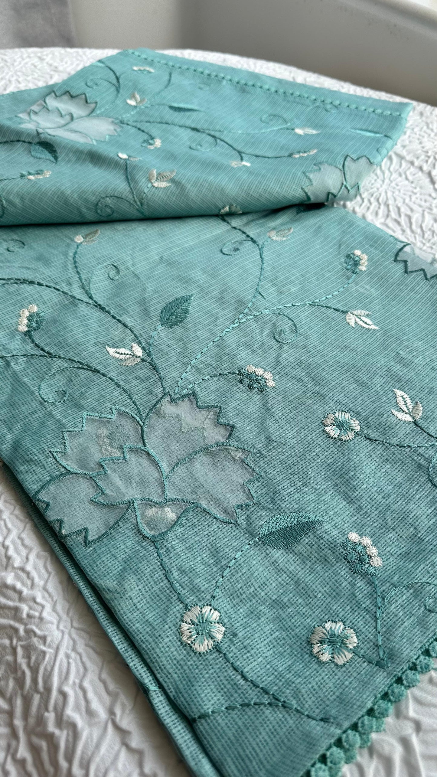Beautiful sage green kota cotton saree with delicate white and green applique floral work scattered across the fabric, featuring decorative border trim, displayed on white textured background