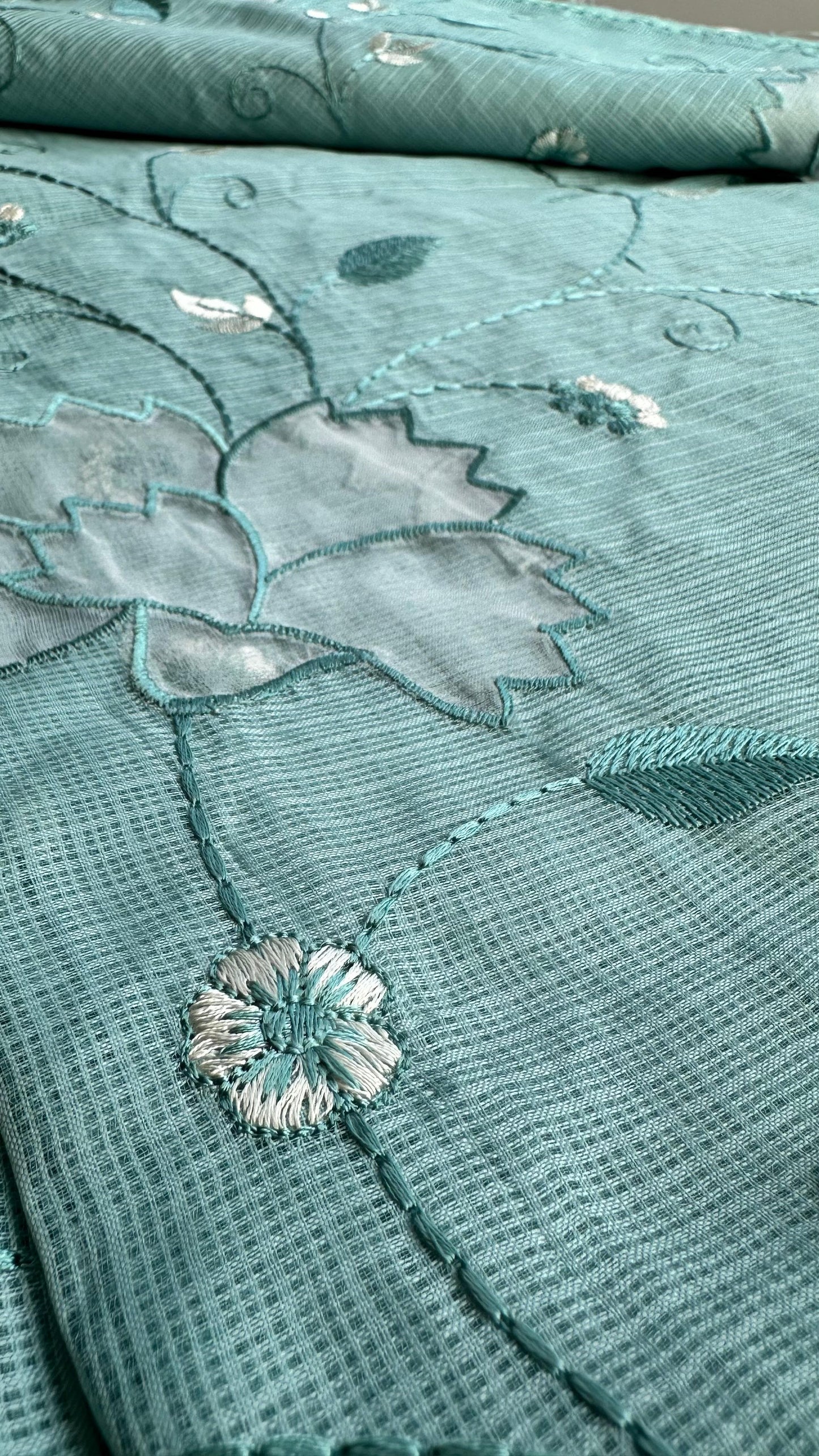Beautiful sage green kota cotton saree with delicate white and green applique floral work scattered across the fabric, featuring decorative border trim, displayed on white textured background