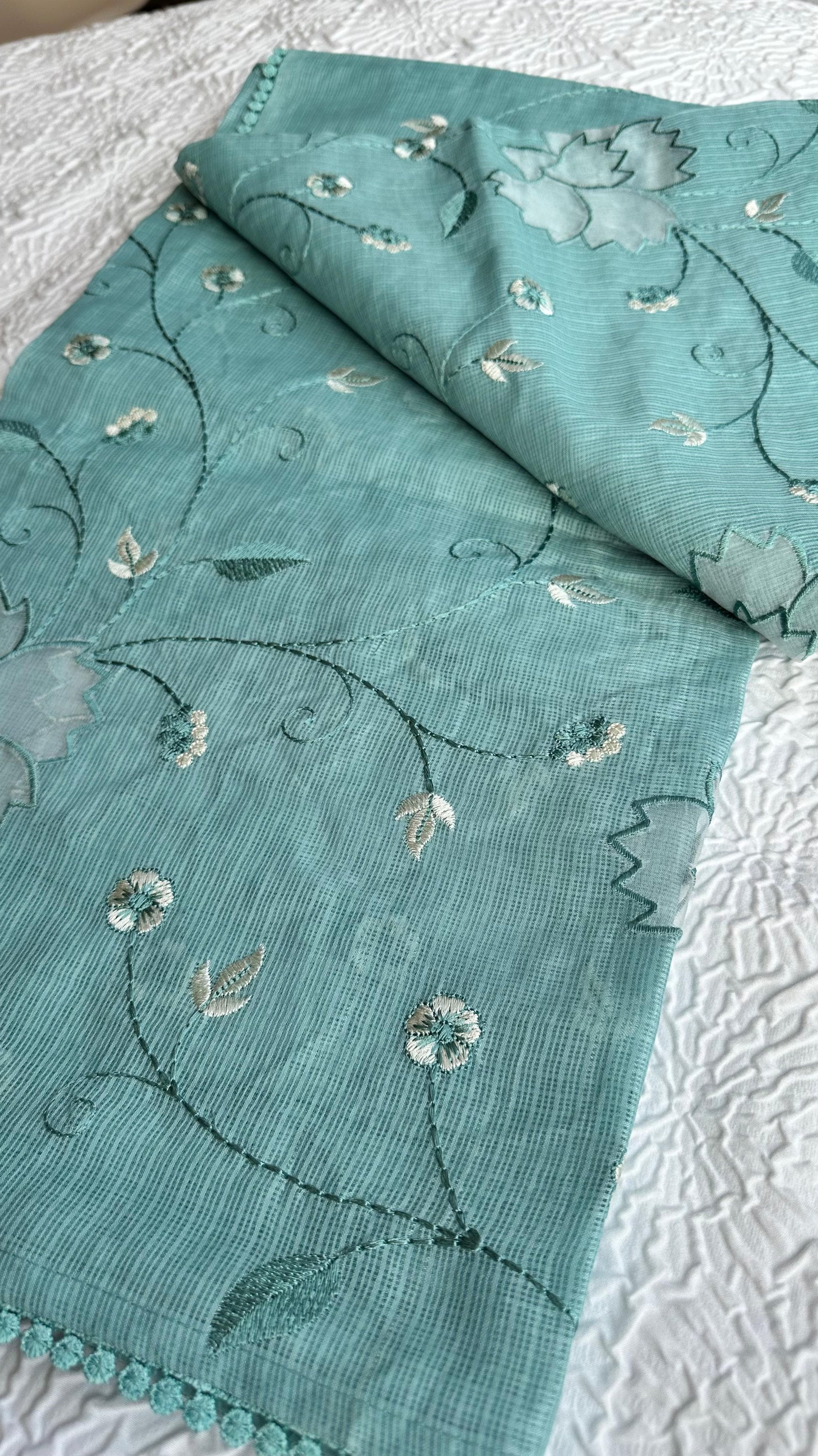 Beautiful sage green kota cotton saree with delicate white and green applique floral work scattered across the fabric, featuring decorative border trim, displayed on white textured background