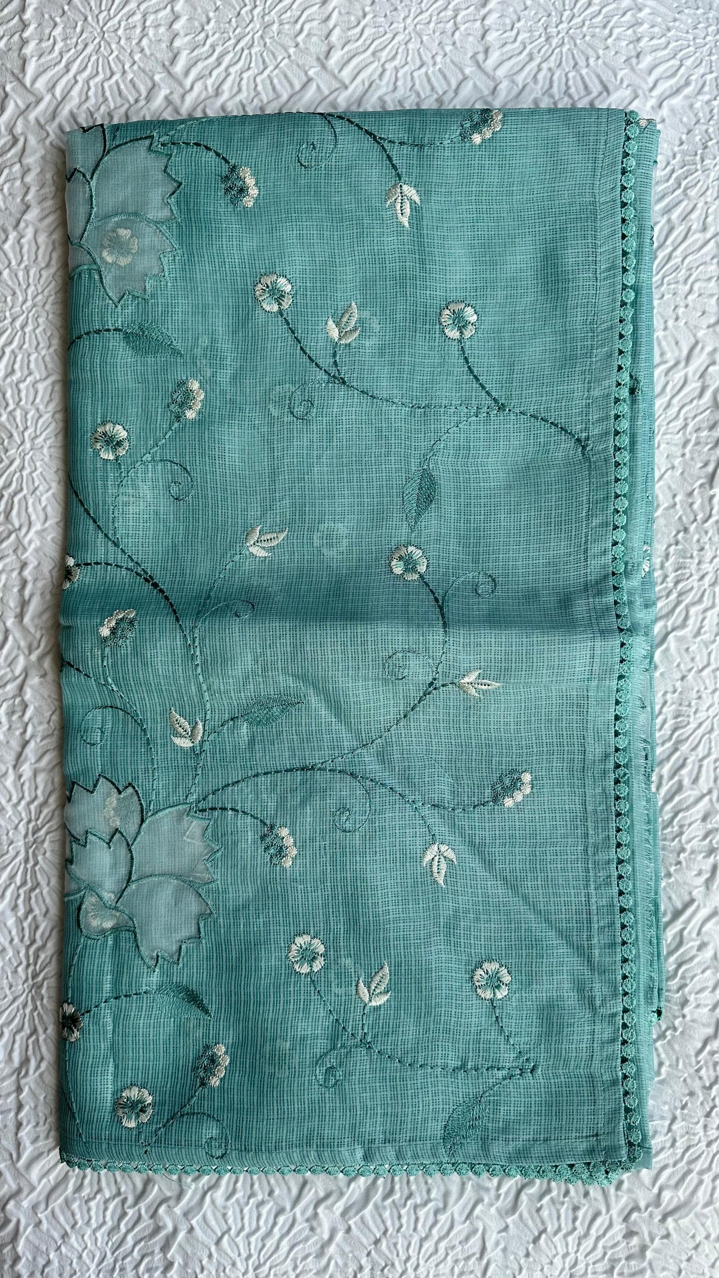Beautiful sage green kota cotton saree with delicate white and green applique floral work scattered across the fabric, featuring decorative border trim, displayed on white textured background