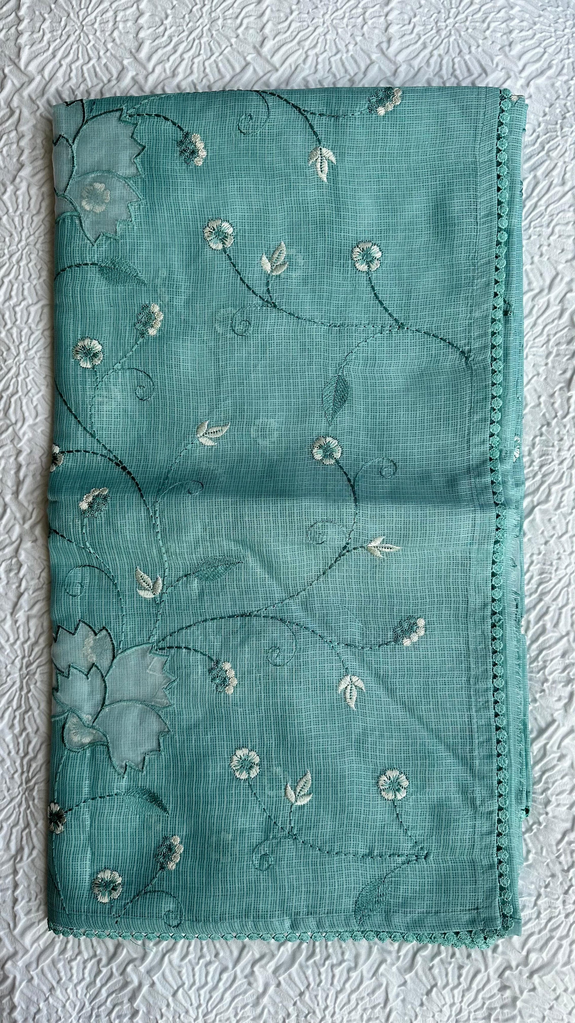 Beautiful sage green kota cotton saree with delicate white and green applique floral work scattered across the fabric, featuring decorative border trim, displayed on white textured background