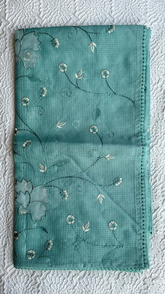 Beautiful sage green kota cotton saree with delicate white and green applique floral work scattered across the fabric, featuring decorative border trim, displayed on white textured background