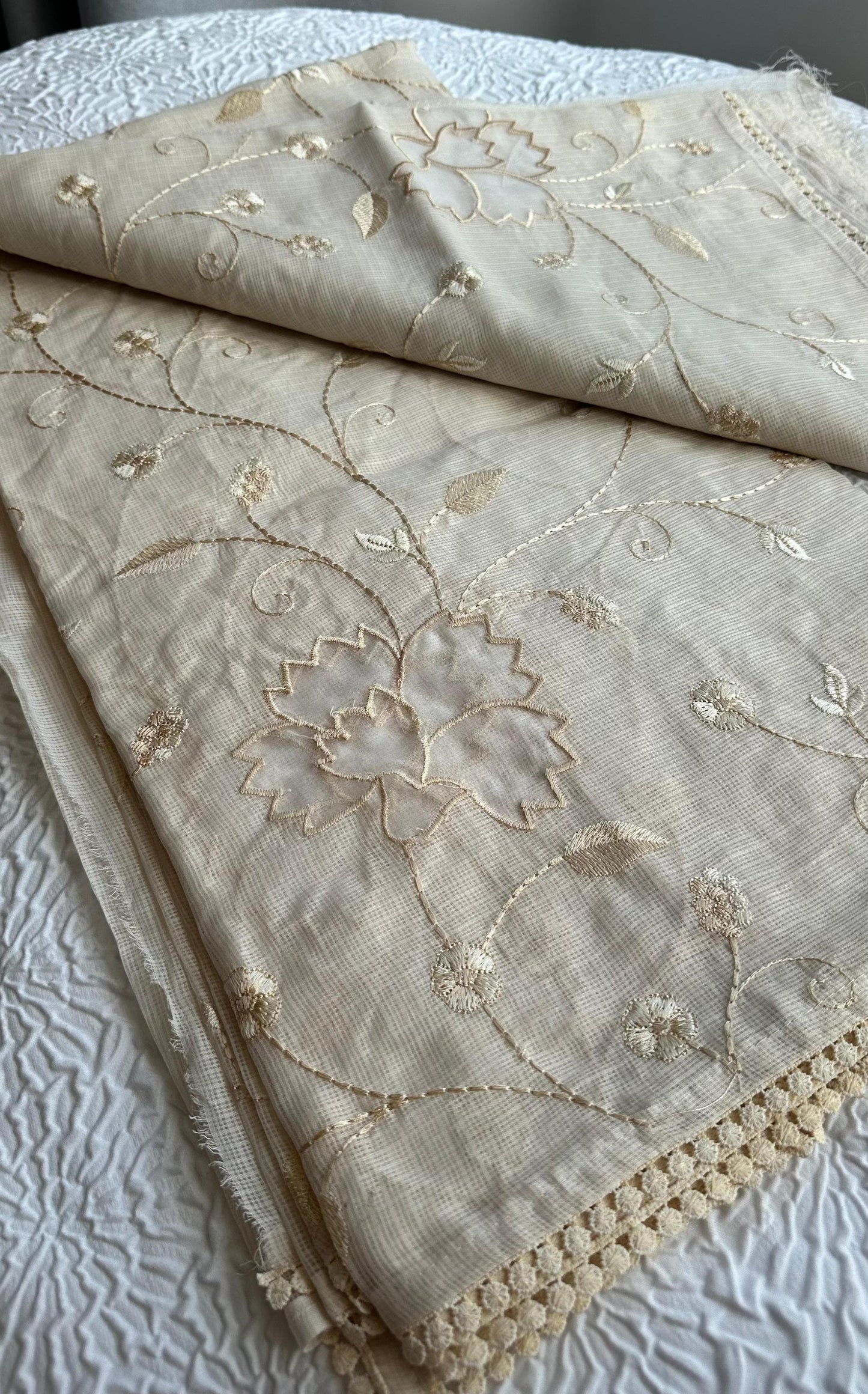 Elegant champagne beige kota cotton saree with delicate gold applique work featuring floral vine patterns and lotus motifs, beautifully folded and displayed on white textured background