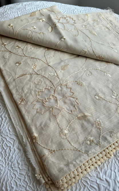 Elegant champagne beige kota cotton saree with delicate gold applique work featuring floral vine patterns and lotus motifs, beautifully folded and displayed on white textured background