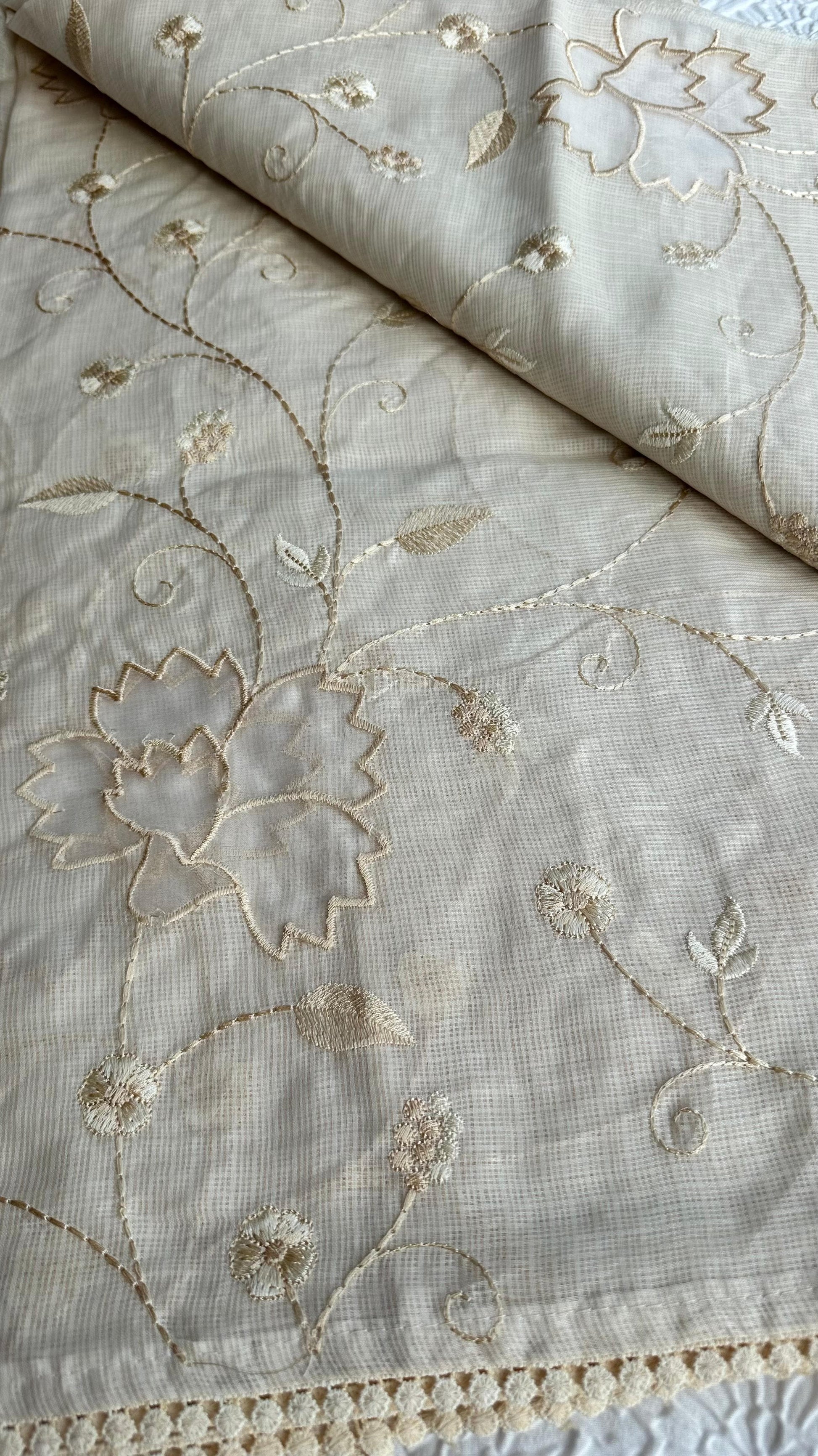 Elegant champagne beige kota cotton saree with delicate gold applique work featuring floral vine patterns and lotus motifs, beautifully folded and displayed on white textured background