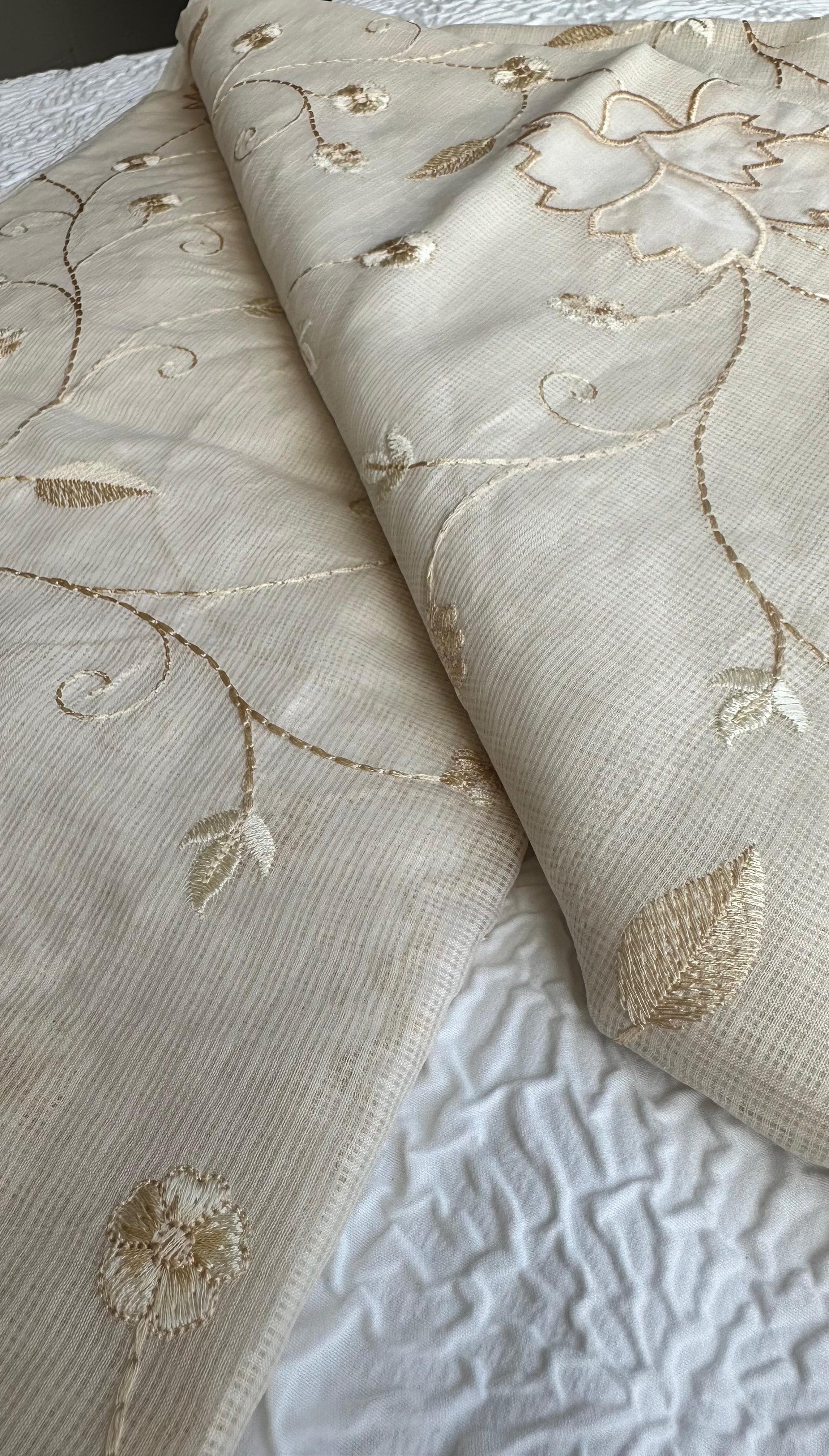 Elegant champagne beige kota cotton saree with delicate gold applique work featuring floral vine patterns and lotus motifs, beautifully folded and displayed on white textured background