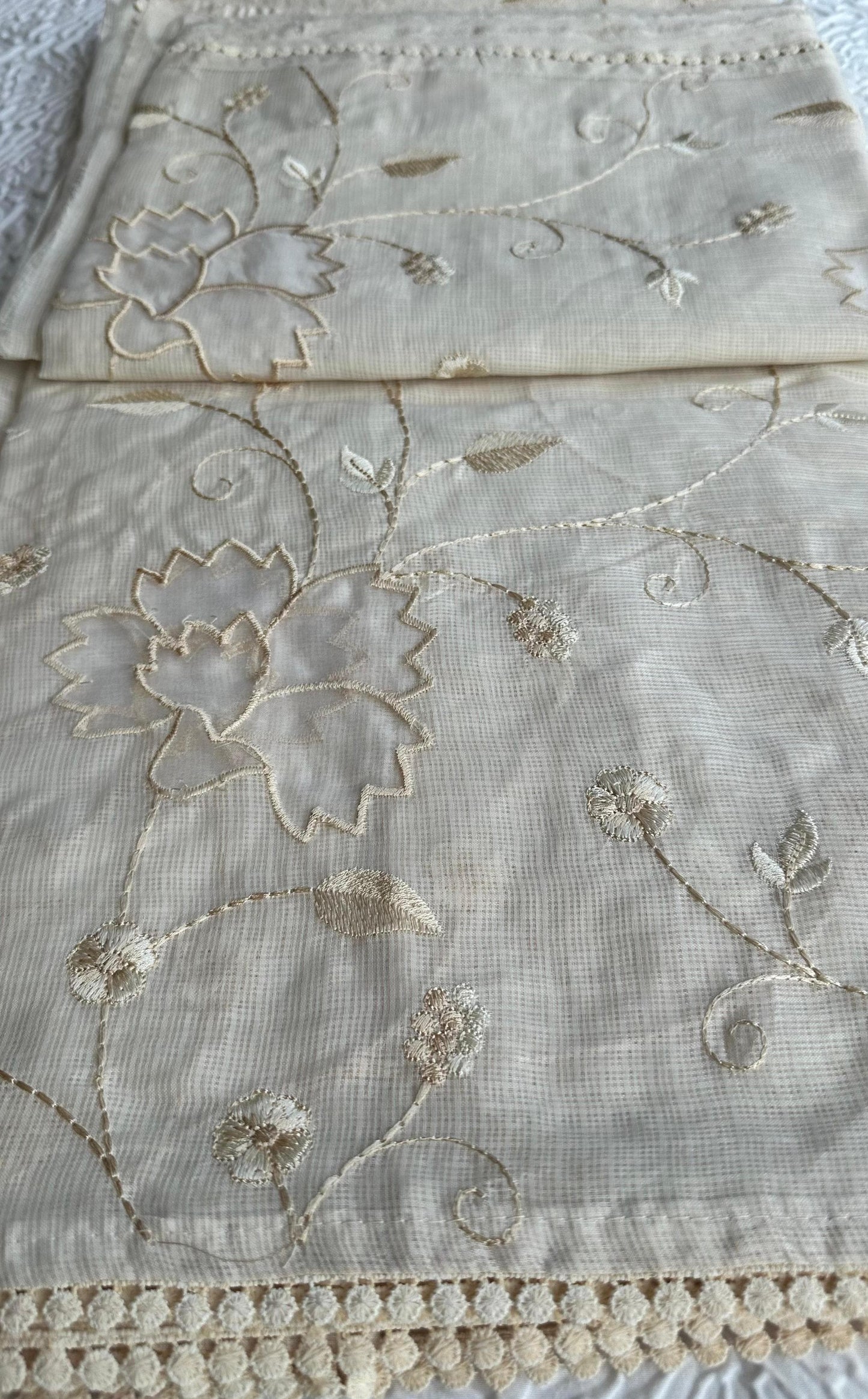 Elegant champagne beige kota cotton saree with delicate gold applique work featuring floral vine patterns and lotus motifs, beautifully folded and displayed on white textured background