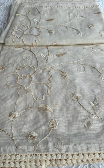 Elegant champagne beige kota cotton saree with delicate gold applique work featuring floral vine patterns and lotus motifs, beautifully folded and displayed on white textured background