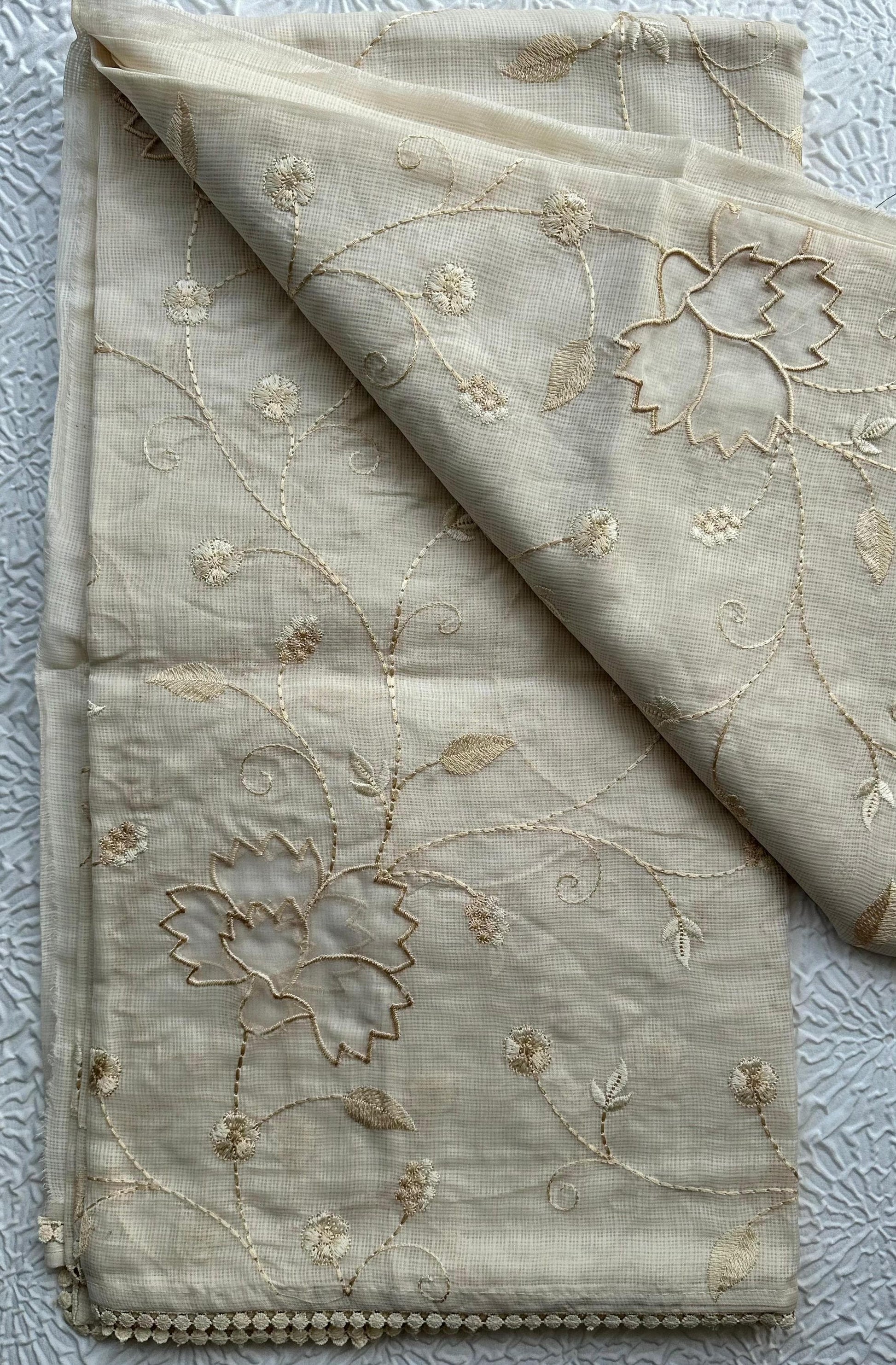 Elegant champagne beige kota cotton saree with delicate gold applique work featuring floral vine patterns and lotus motifs, beautifully folded and displayed on white textured background
