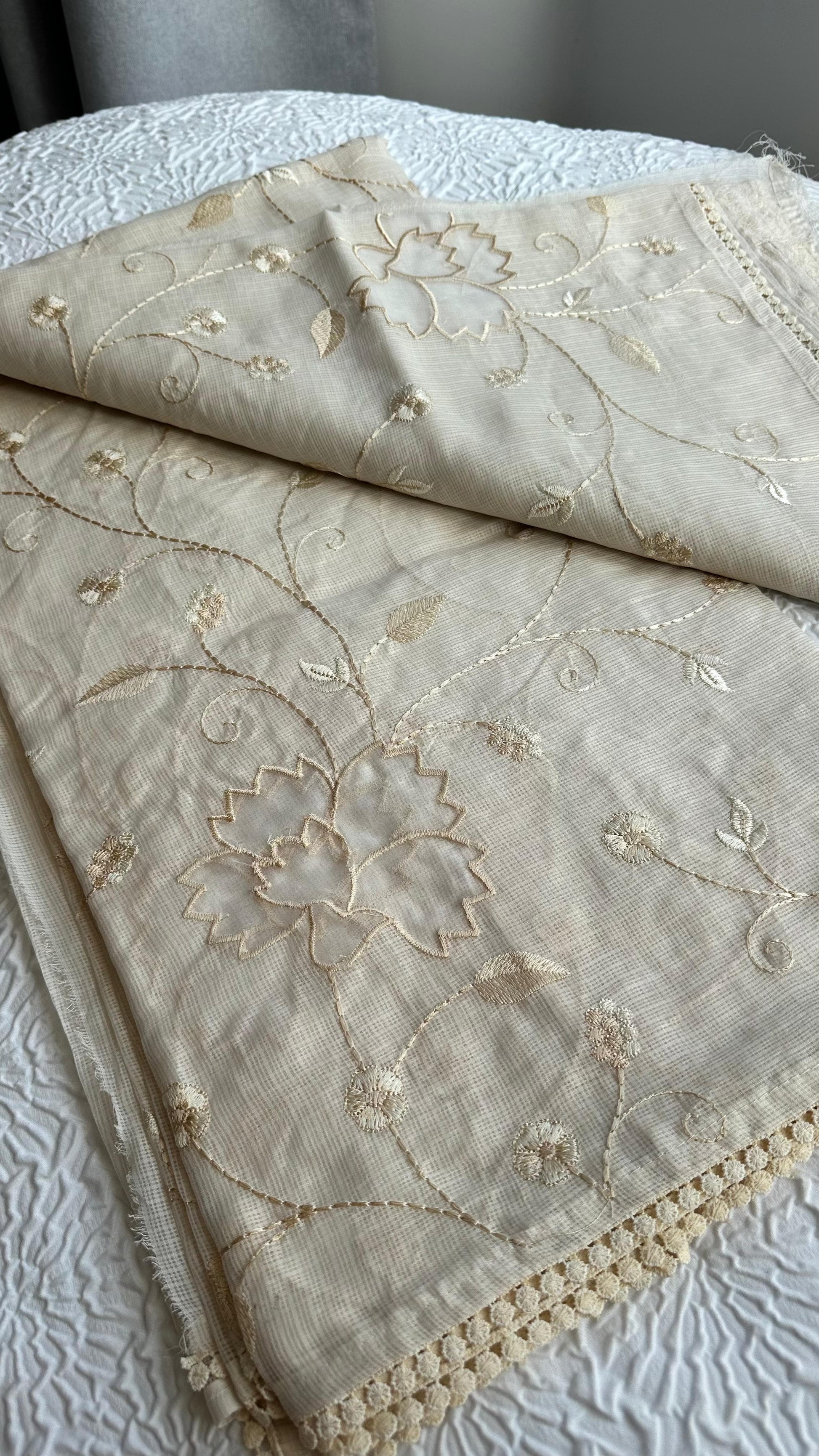 Elegant champagne beige kota cotton saree with delicate gold applique work featuring floral vine patterns and lotus motifs, beautifully folded and displayed on white textured background
