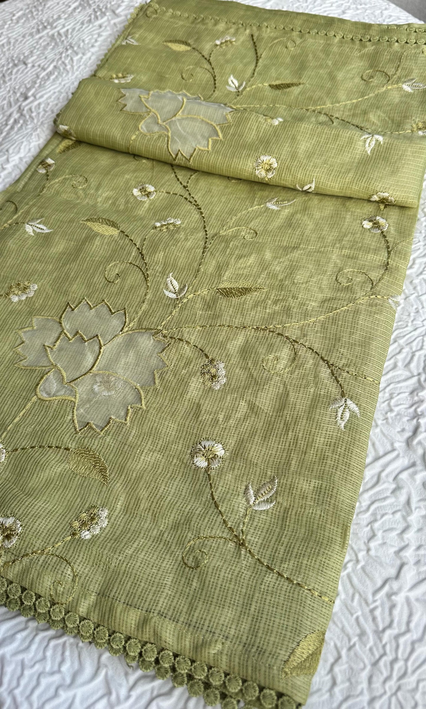 Beautiful olive green kota cotton saree with delicate lotus applique work and floral embroidery scattered across the fabric, featuring decorative scalloped border, displayed on white textured background
