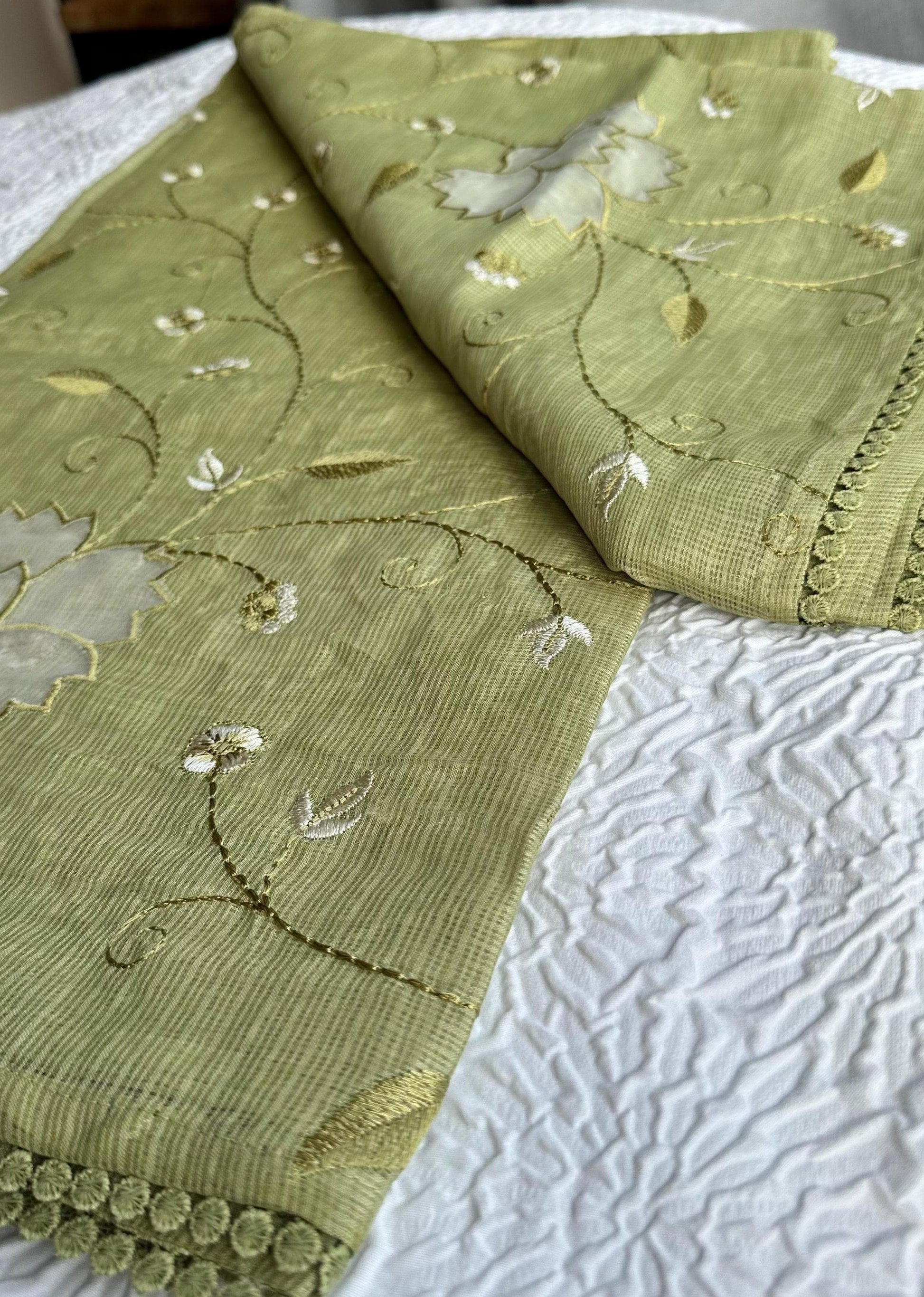 Beautiful olive green kota cotton saree with delicate lotus applique work and floral embroidery scattered across the fabric, featuring decorative scalloped border, displayed on white textured background