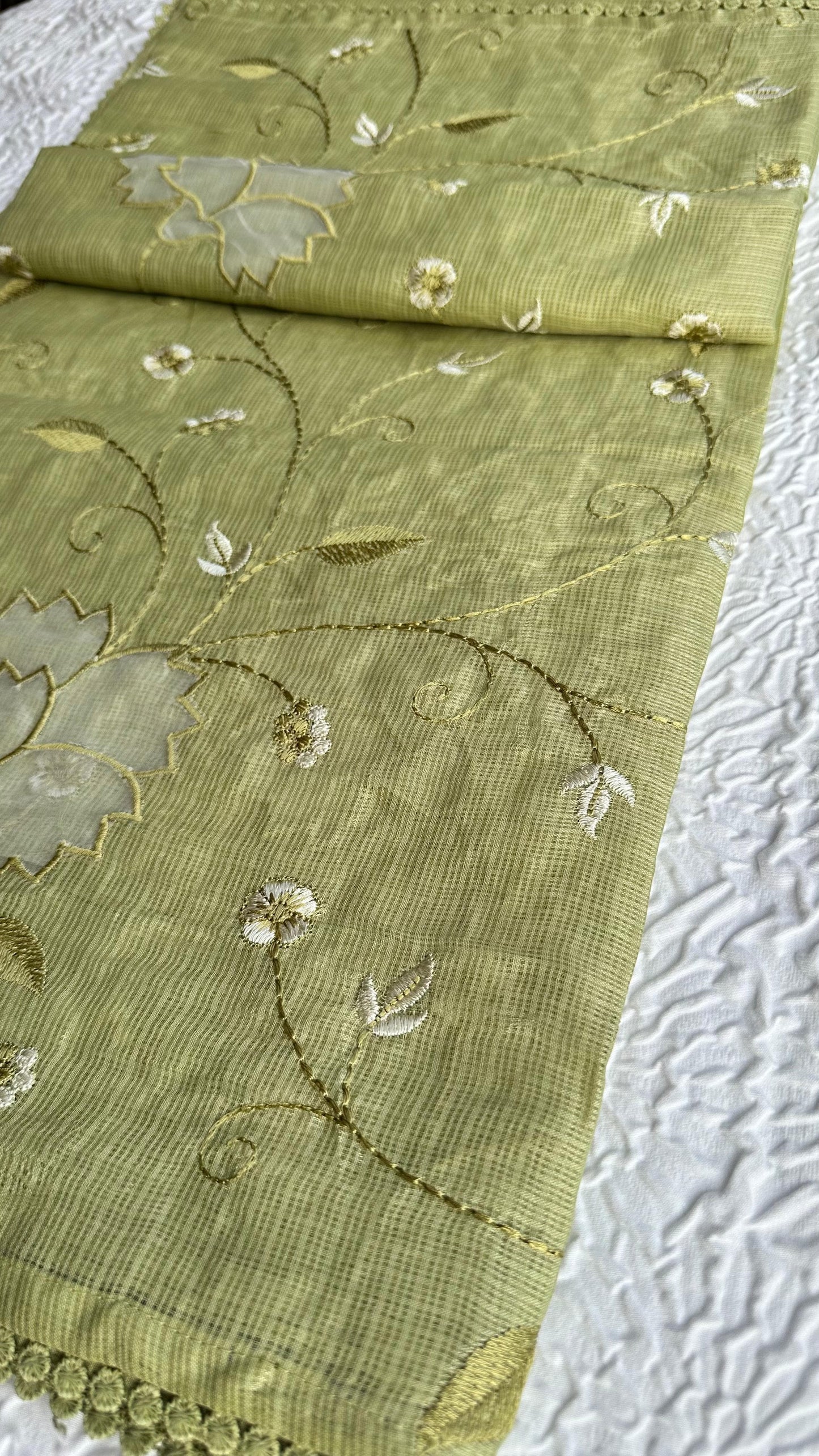 Beautiful olive green kota cotton saree with delicate lotus applique work and floral embroidery scattered across the fabric, featuring decorative scalloped border, displayed on white textured background