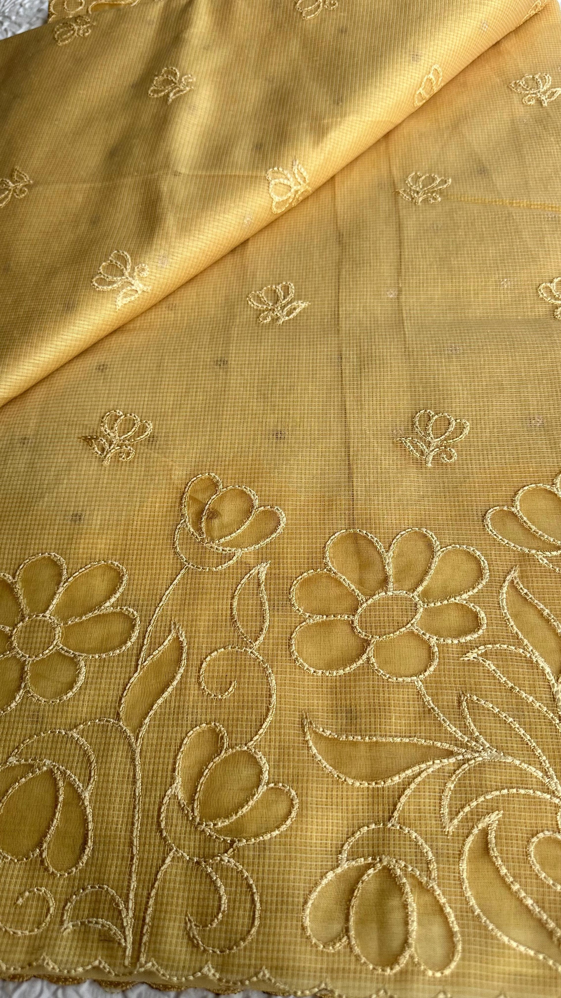 Radiant golden yellow kota cotton saree with beautiful floral applique work featuring delicate flower motifs and vine patterns with scalloped border detailing, elegantly displayed on white textured background