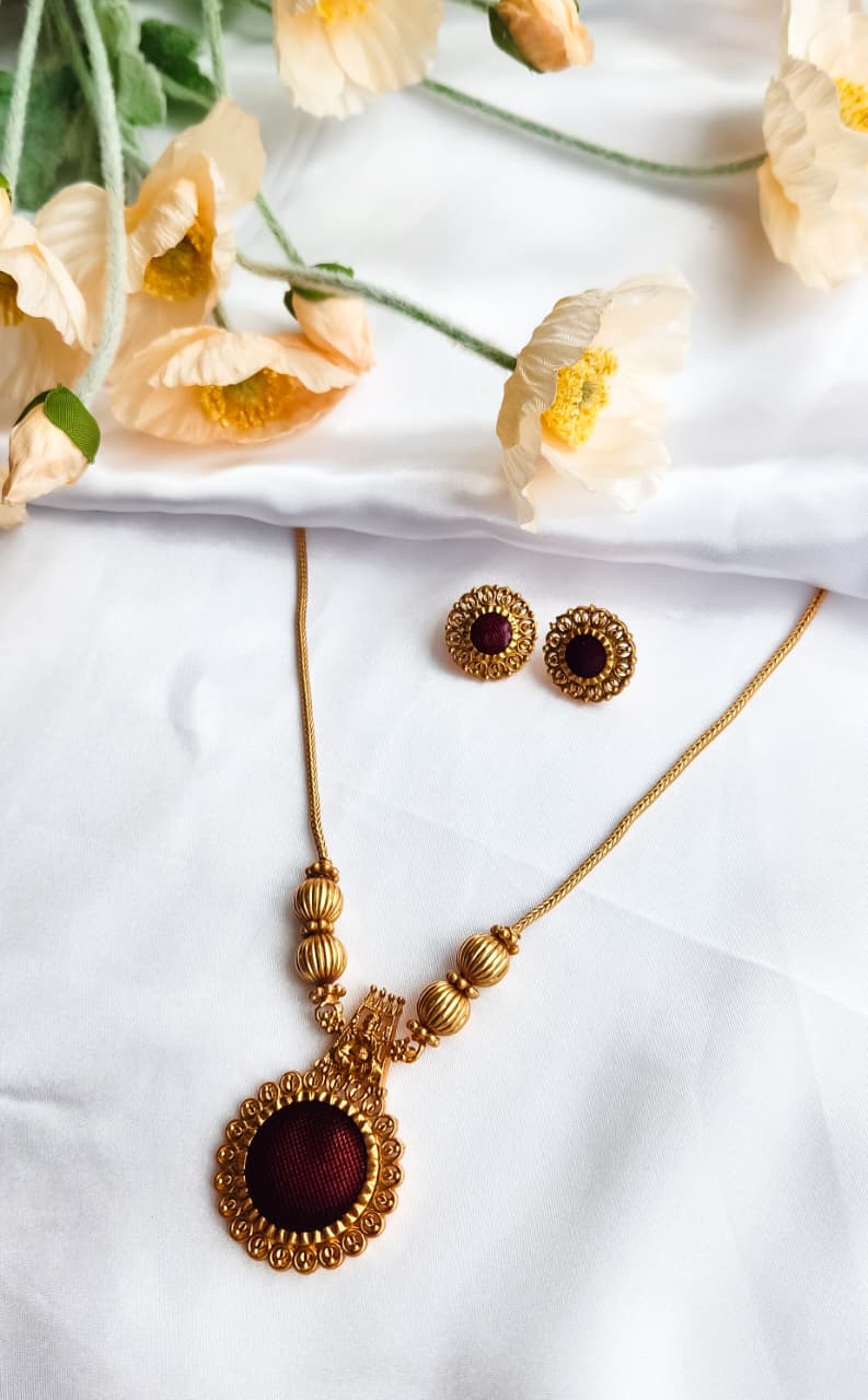 Traditional gold-toned temple jewelry necklace set with maroon circular pendant and matching earrings, displayed on white silk with flowers.
