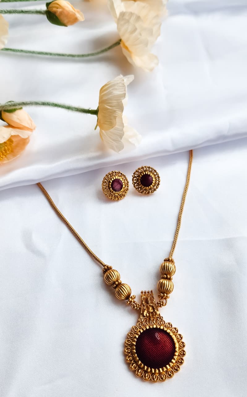 Traditional gold-toned temple jewelry necklace set with maroon circular pendant and matching earrings, displayed on white silk with flowers.