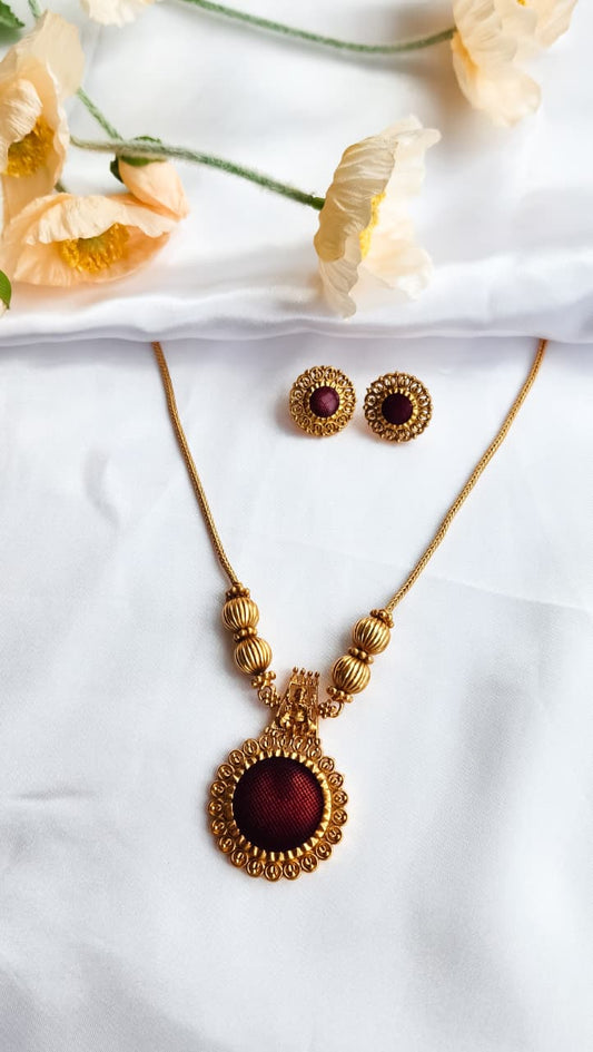 Traditional gold-toned temple jewelry necklace set with maroon circular pendant and matching earrings, displayed on white silk with flowers.
