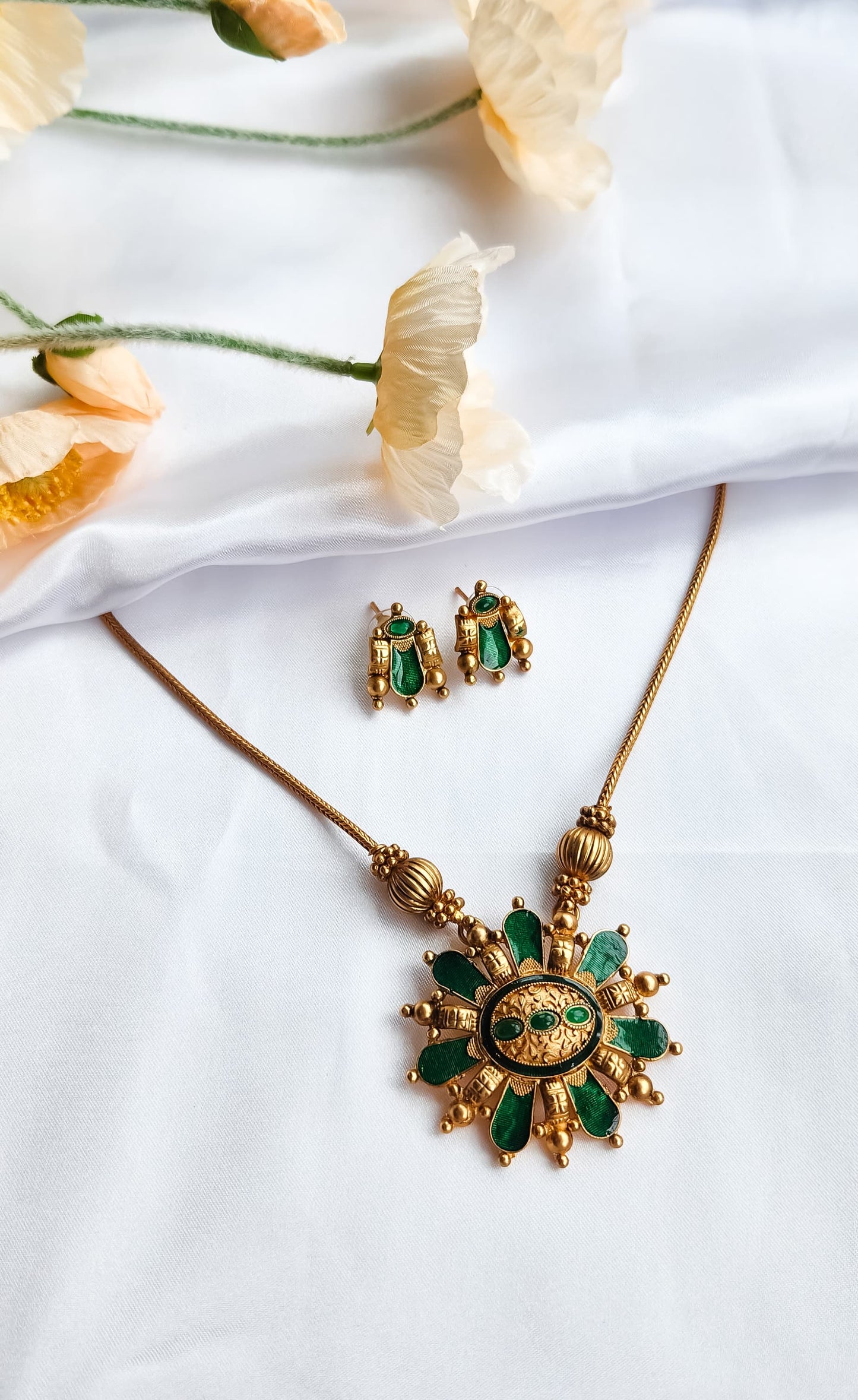 Traditional green and gold sunburst necklace set with matching earrings displayed on white silk cloth with flowers.