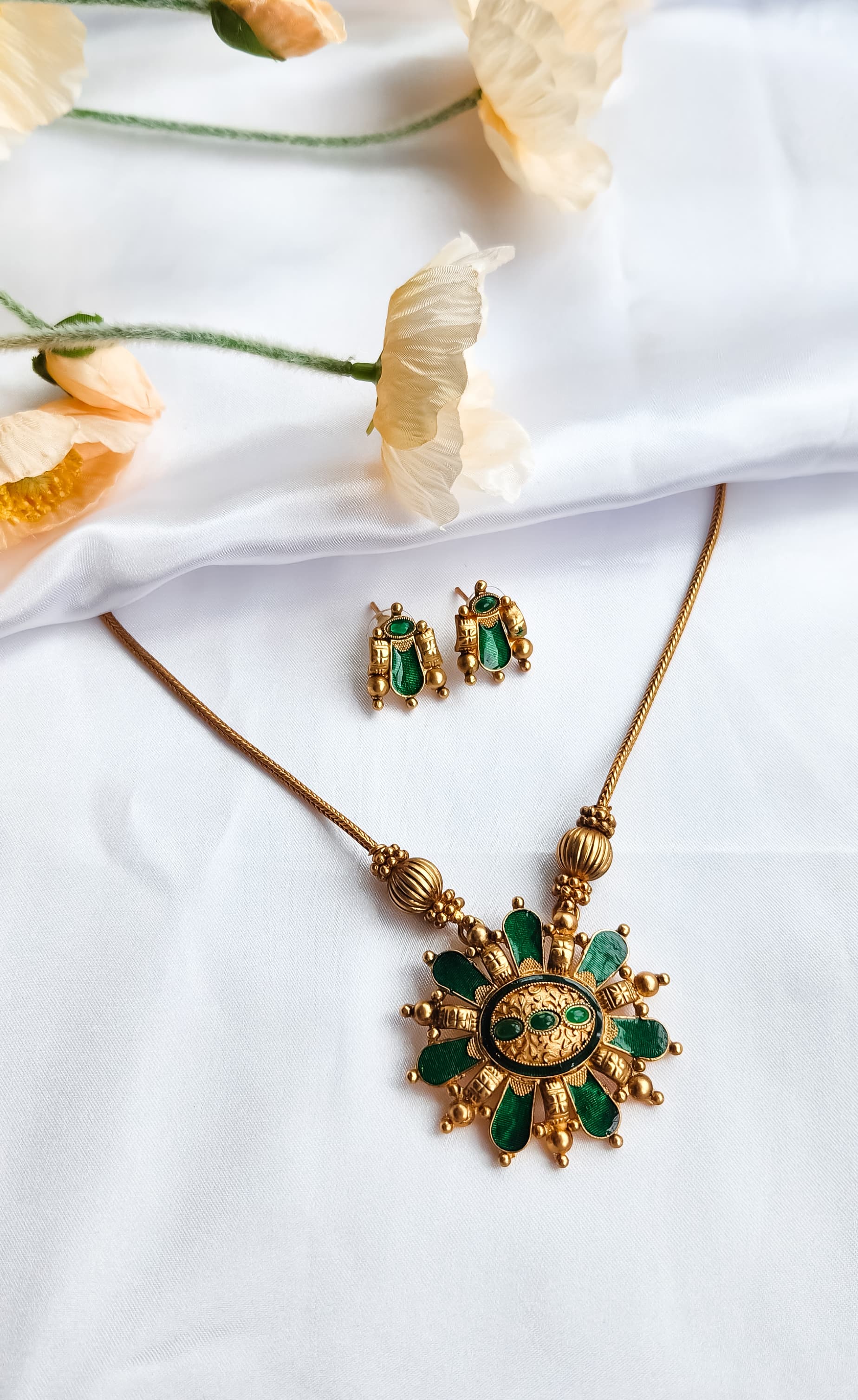 Traditional green and gold sunburst necklace set with matching earrings displayed on white silk cloth with flowers.