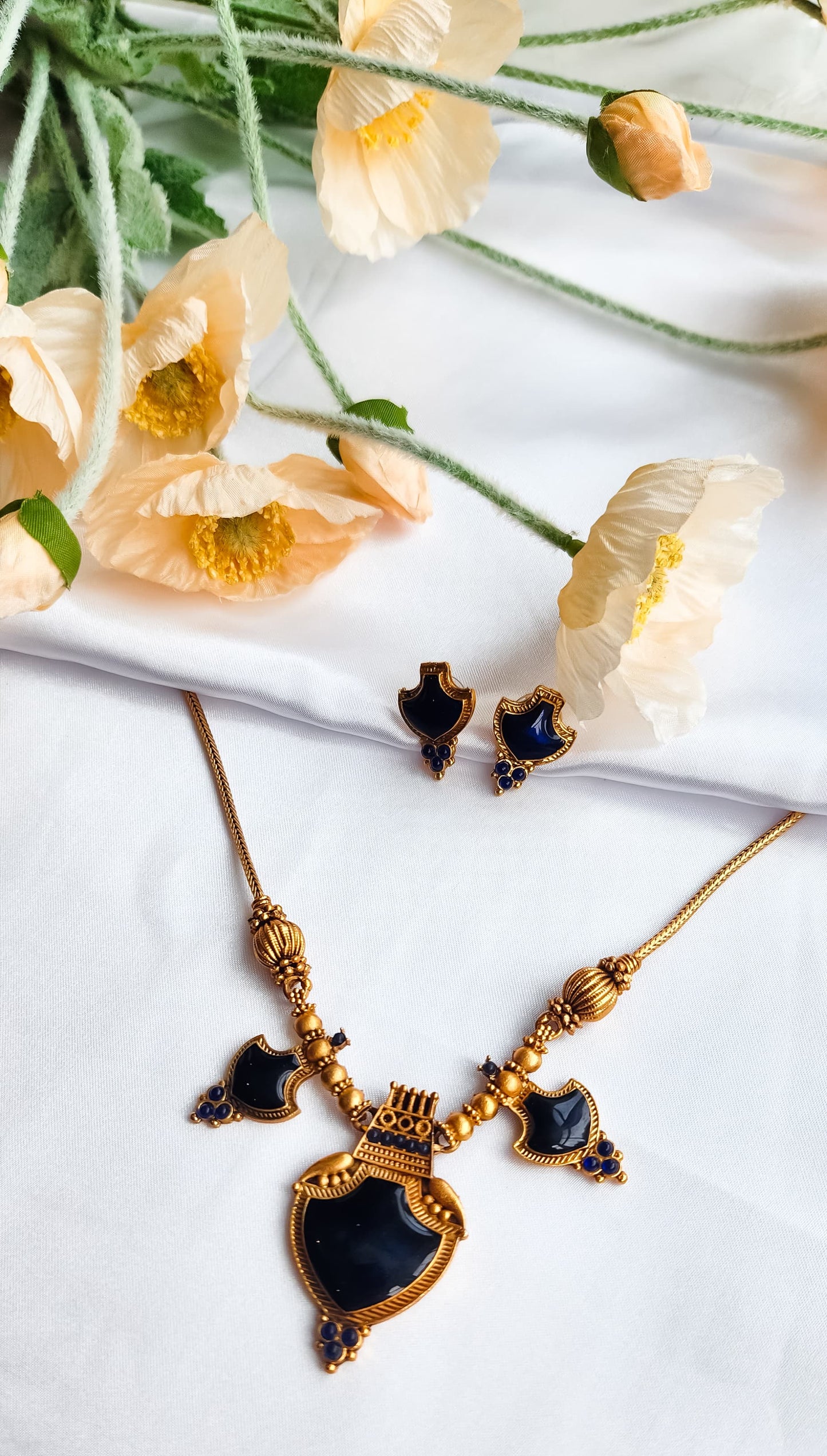 Antique gold necklace set with royal blue stones and shield-shaped pendant, paired with matching earrings, displayed on white fabric with flowers.