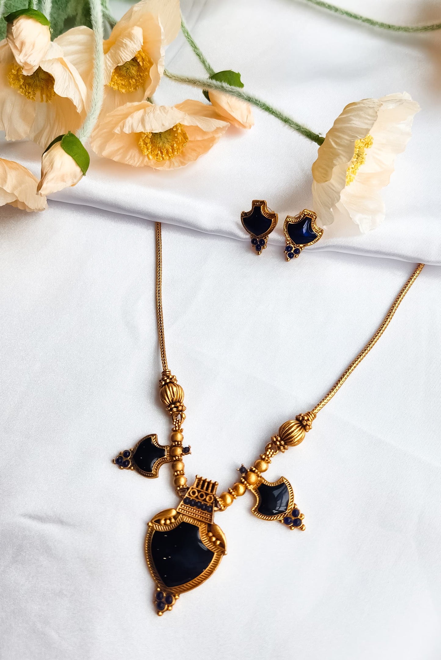 Antique gold necklace set with royal blue stones and shield-shaped pendant, paired with matching earrings, displayed on white fabric with flowers.