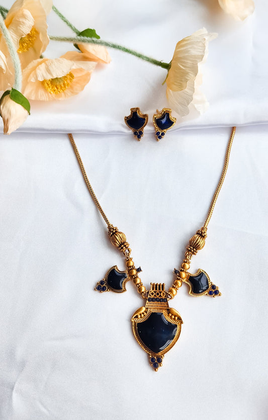 Antique gold necklace set with royal blue stones and shield-shaped pendant, paired with matching earrings, displayed on white fabric with flowers.