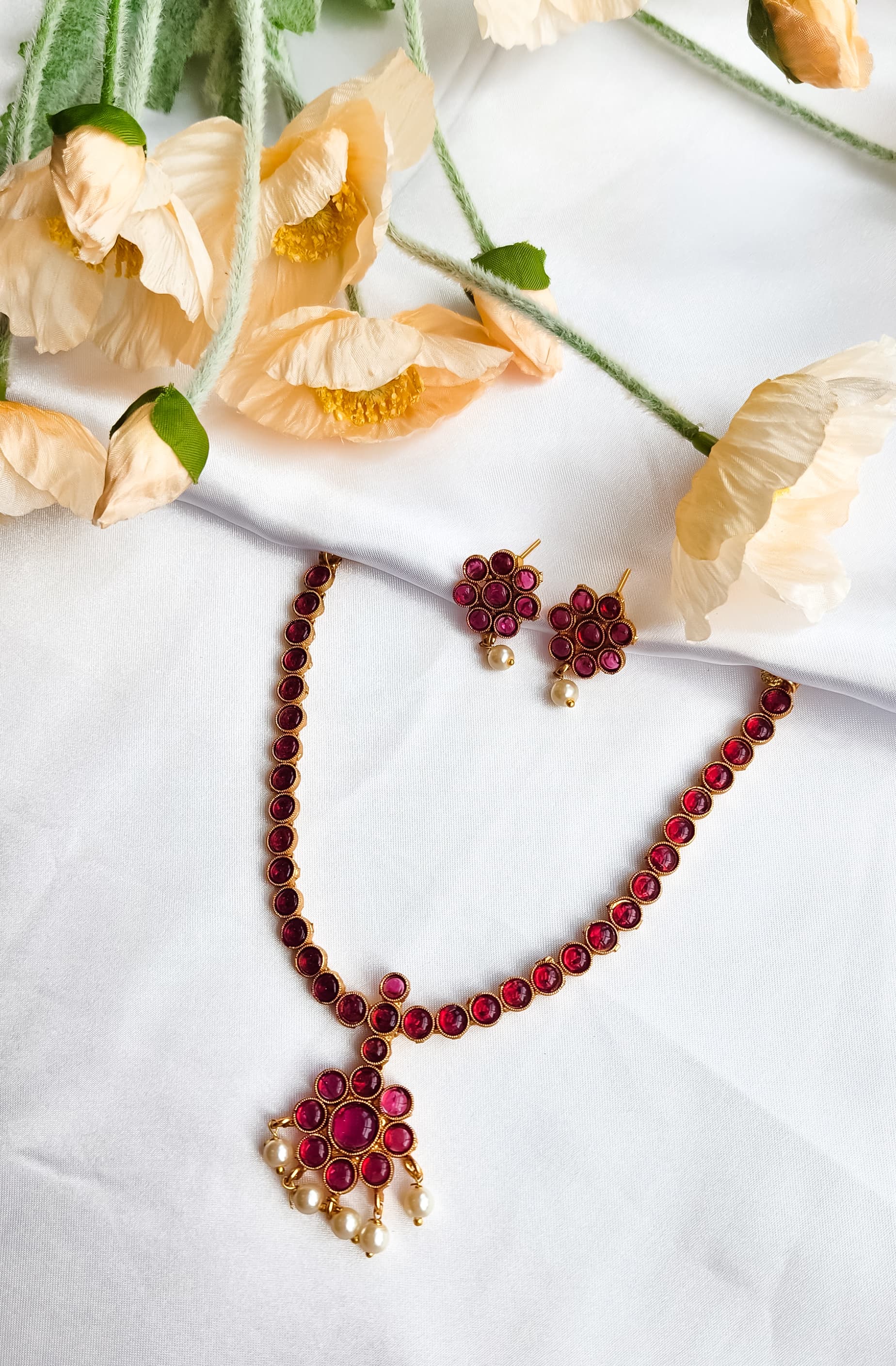 Traditional red stone necklace set with floral pendant, pearl drop accents, and matching red stone earrings displayed on white fabric with flowers.