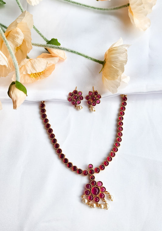 Traditional red stone necklace set with floral pendant, pearl drop accents, and matching red stone earrings displayed on white fabric with flowers.