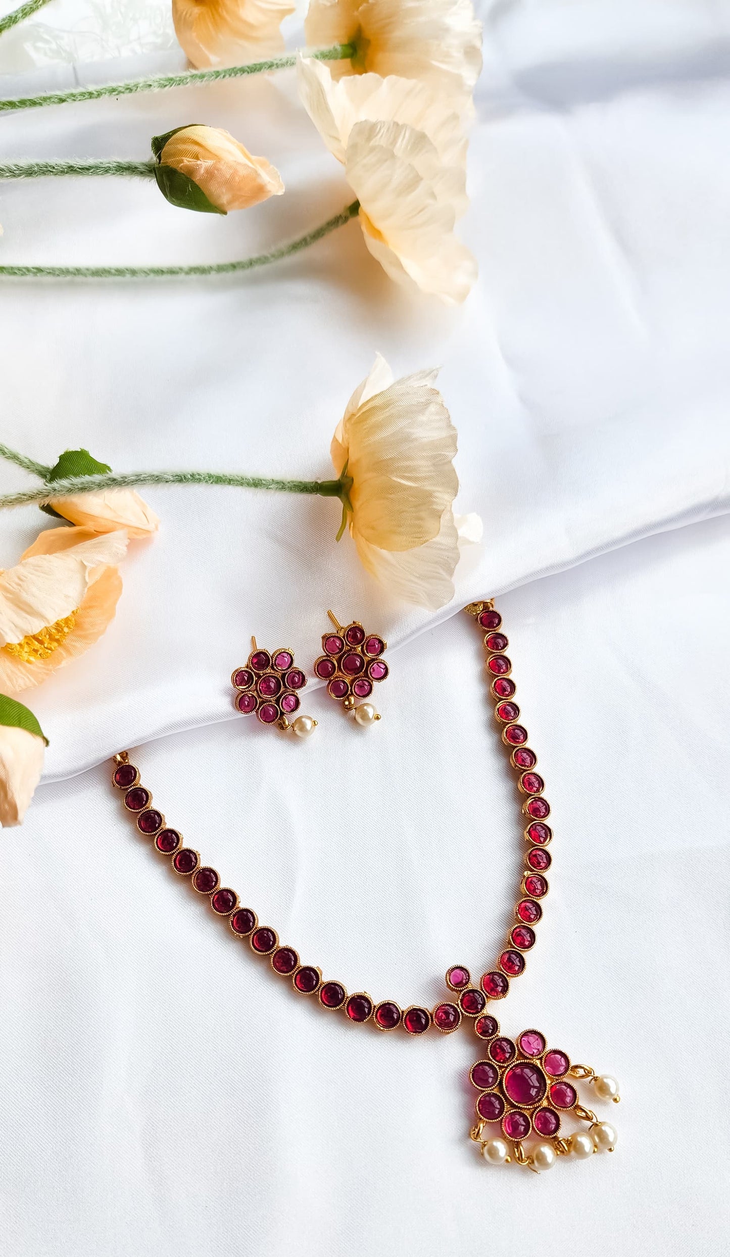 Traditional red stone necklace set with floral pendant, pearl drop accents, and matching red stone earrings displayed on white fabric with flowers.