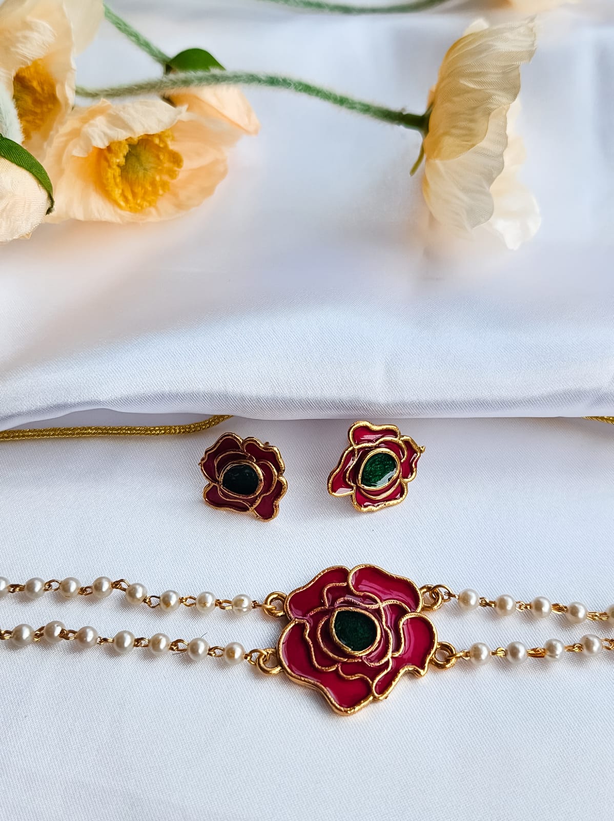 A rose-inspired jewelry set with red and green enamel detailing, featuring a pearl-strung bracelet and matching rose motif earrings, displayed on a white background with flowers.