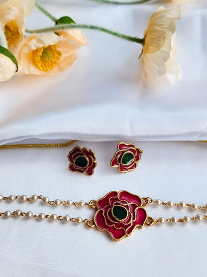 A rose-inspired jewelry set with red and green enamel detailing, featuring a pearl-strung bracelet and matching rose motif earrings, displayed on a white background with flowers.