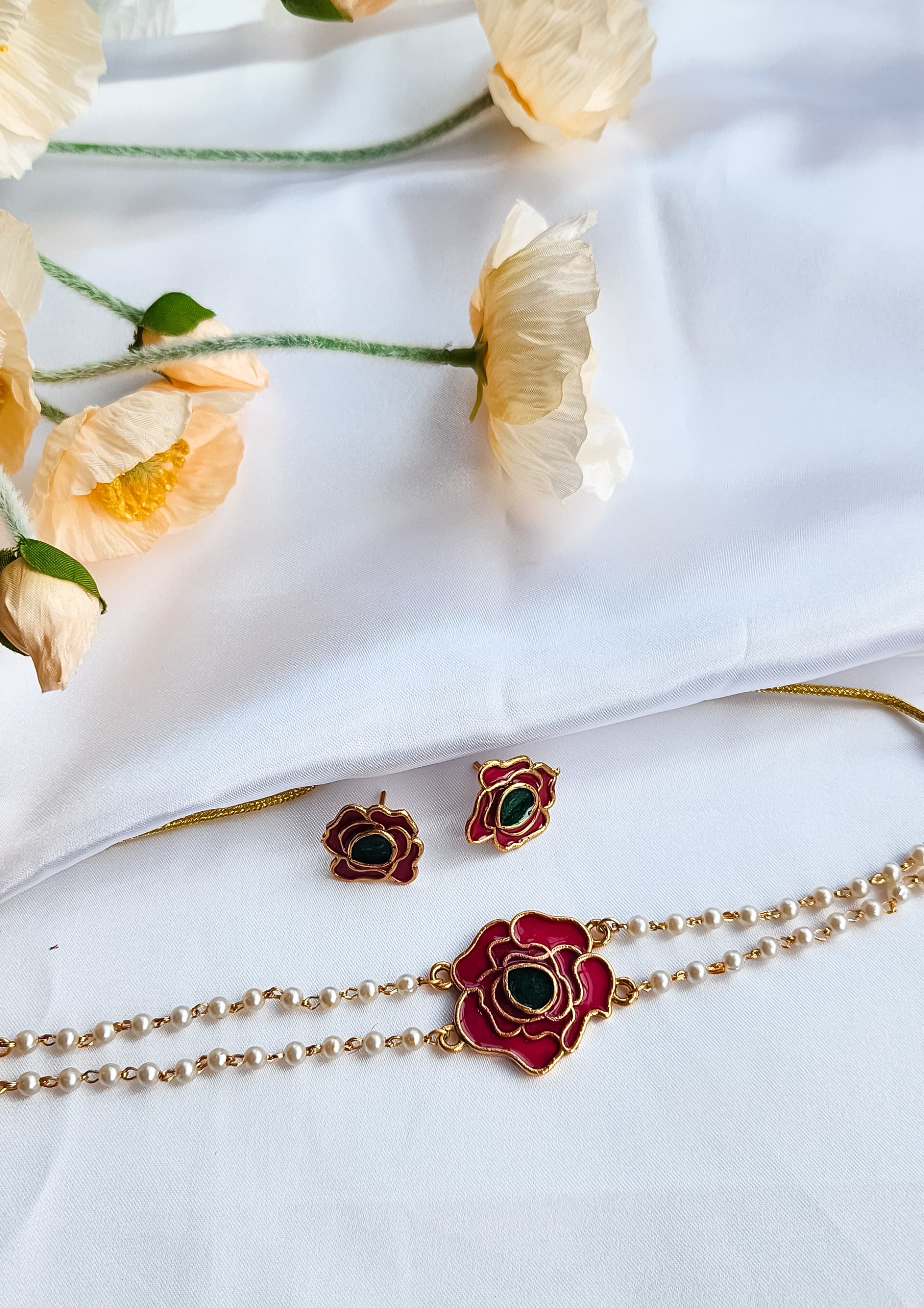 A rose-inspired jewelry set with red and green enamel detailing, featuring a pearl-strung bracelet and matching rose motif earrings, displayed on a white background with flowers.