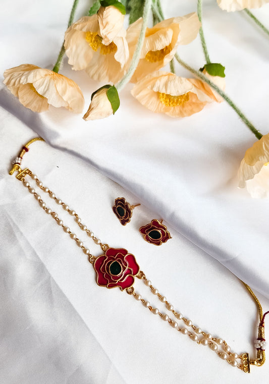 A rose-inspired jewelry set with red and green enamel detailing, featuring a pearl-strung bracelet and matching rose motif earrings, displayed on a white background with flowers.