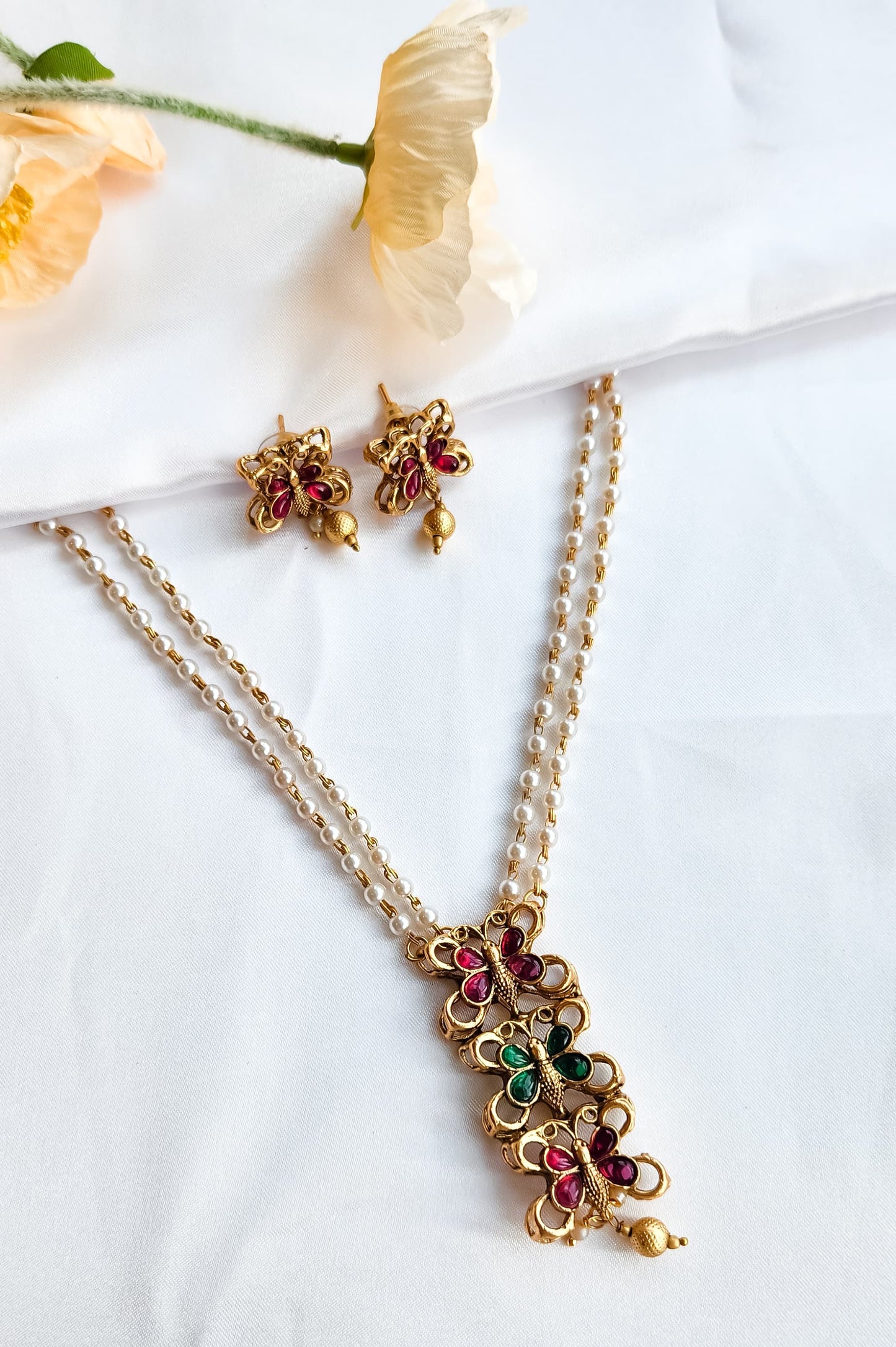 Traditional butterfly design pearl mala set with ruby-pink and emerald-green stones and matching earrings displayed on white fabric with flowers.