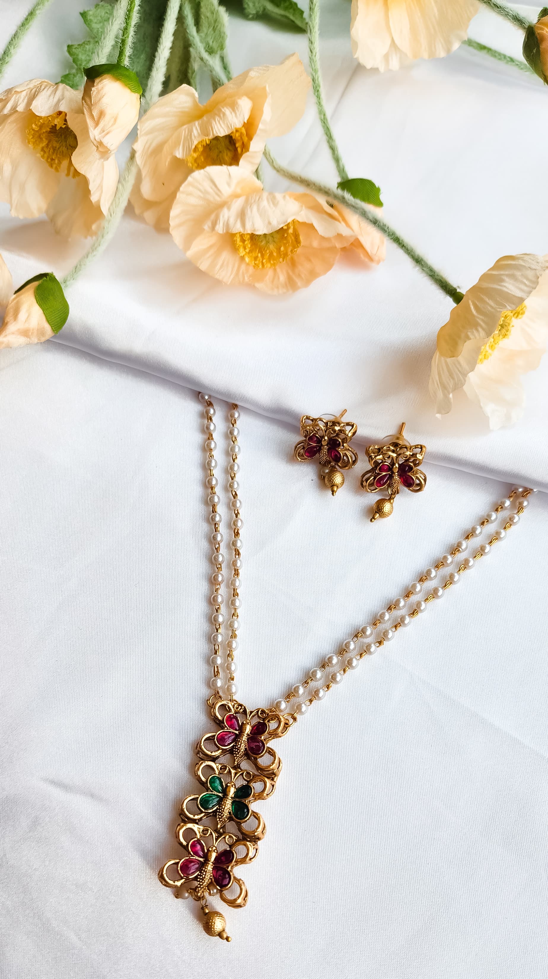 Traditional butterfly design pearl mala set with ruby-pink and emerald-green stones and matching earrings displayed on white fabric with flowers.