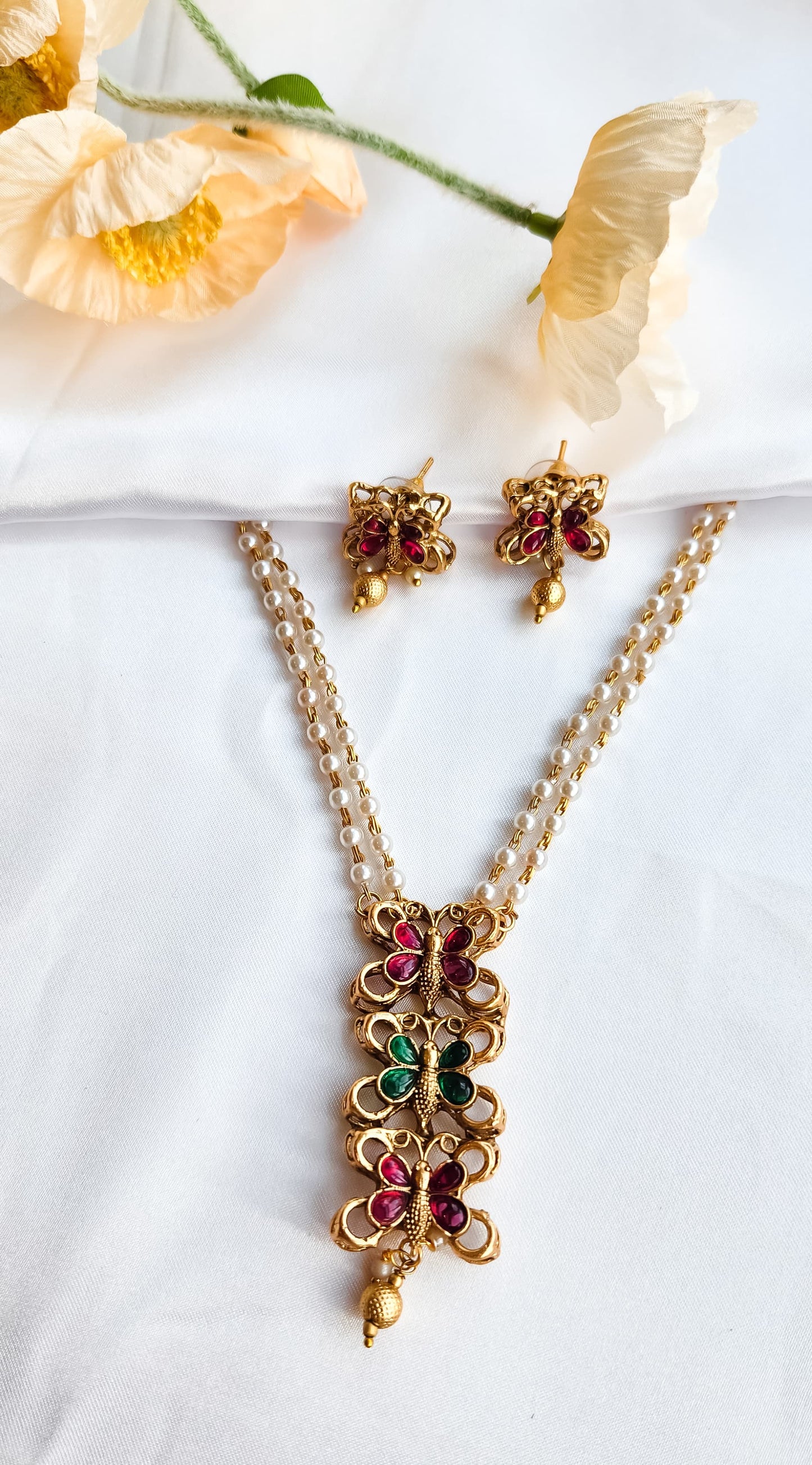 Traditional butterfly design pearl mala set with ruby-pink and emerald-green stones and matching earrings displayed on white fabric with flowers.