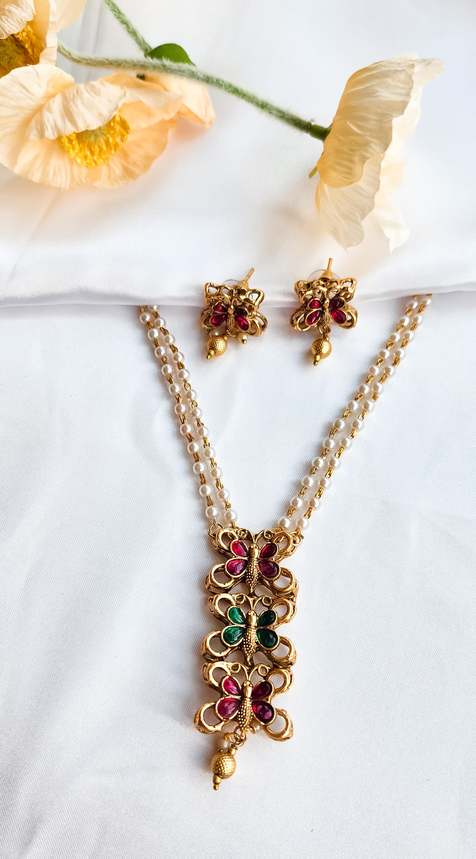 Traditional butterfly design pearl mala set with ruby-pink and emerald-green stones and matching earrings displayed on white fabric with flowers.