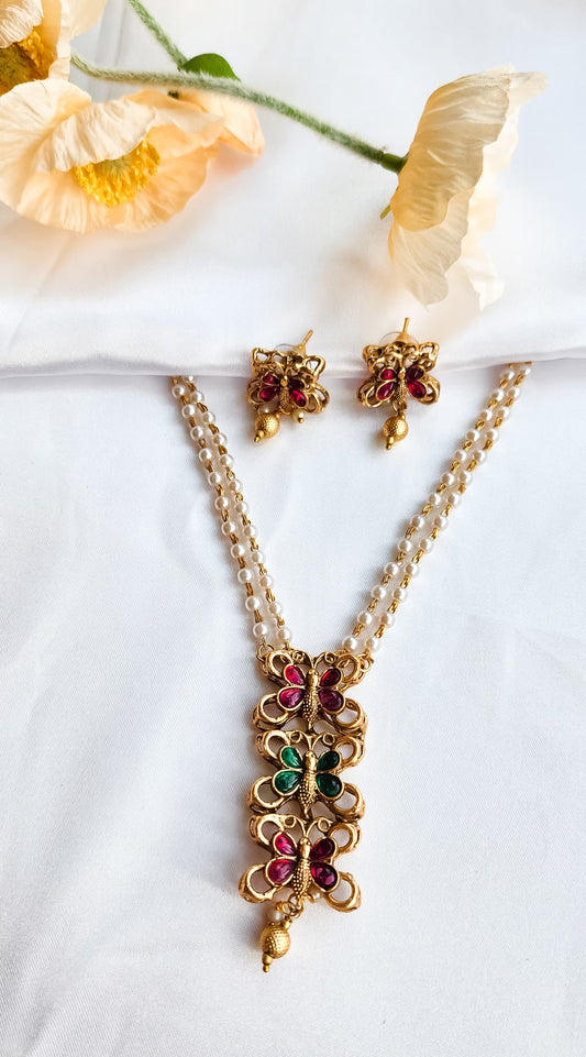 Traditional butterfly design pearl mala set with ruby-pink and emerald-green stones and matching earrings displayed on white fabric with flowers.