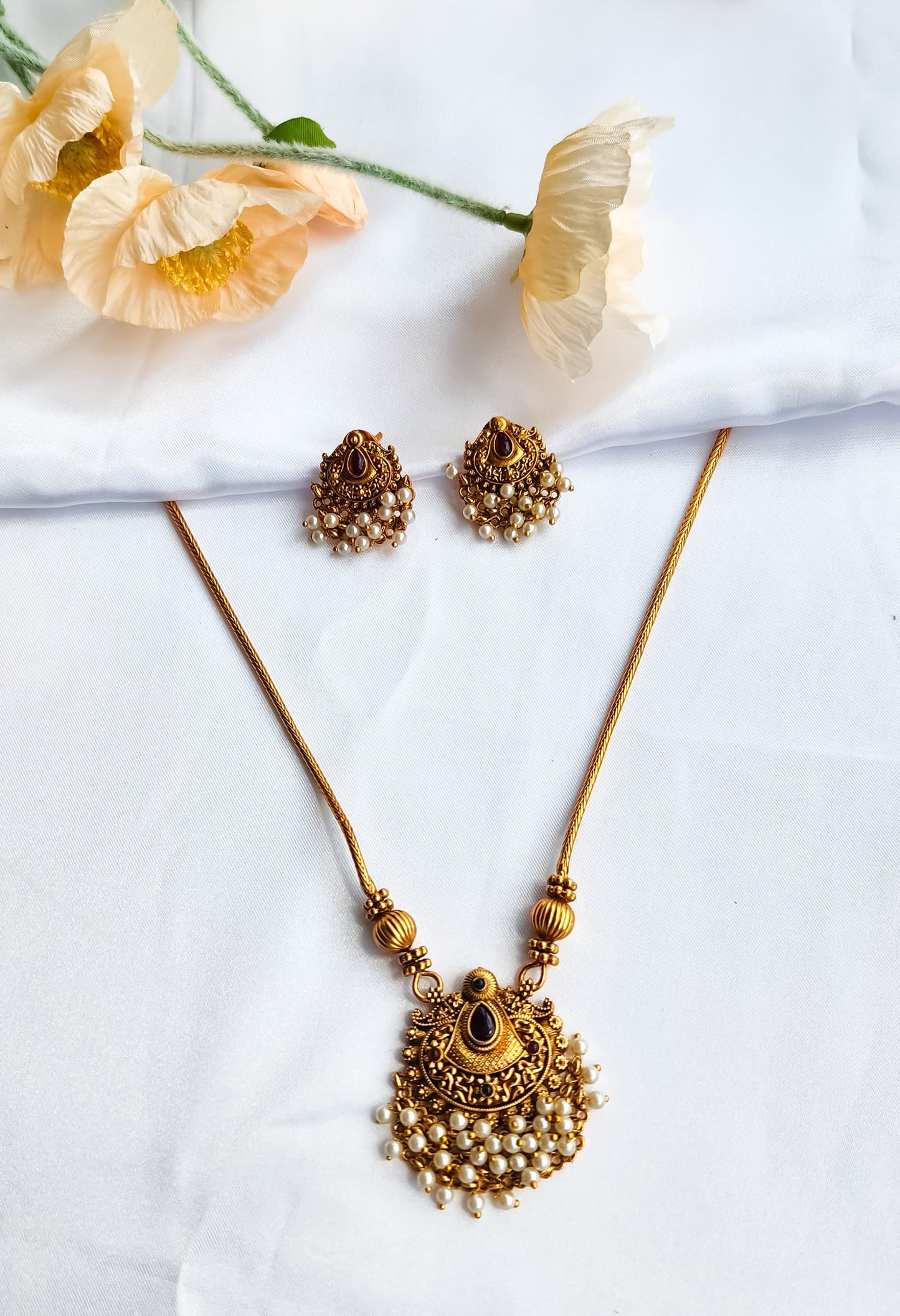 Antique gold temple-style necklace with pearl-detailed pendant and matching earrings on a white background with flowers.