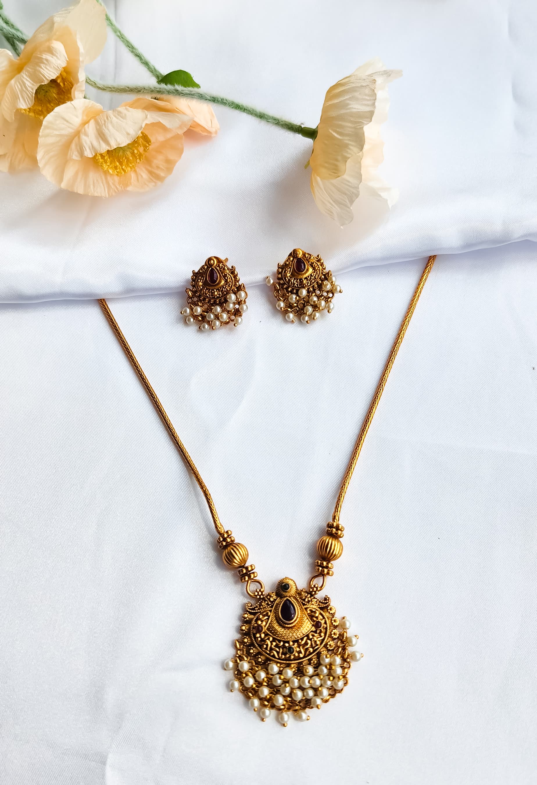 Antique gold temple-style necklace with pearl-detailed pendant and matching earrings on a white background with flowers.