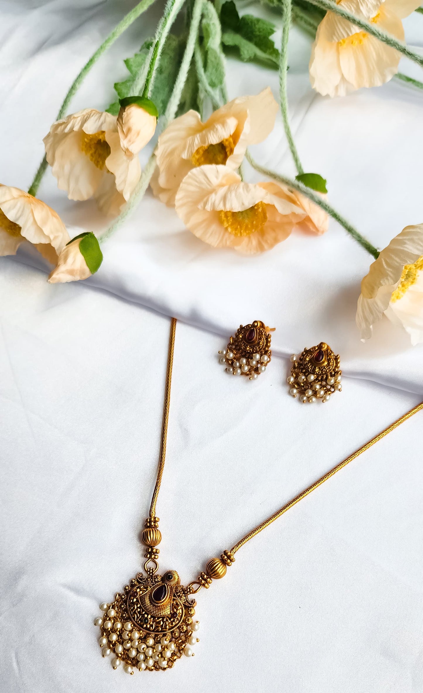 Antique gold temple-style necklace with pearl-detailed pendant and matching earrings on a white background with flowers.