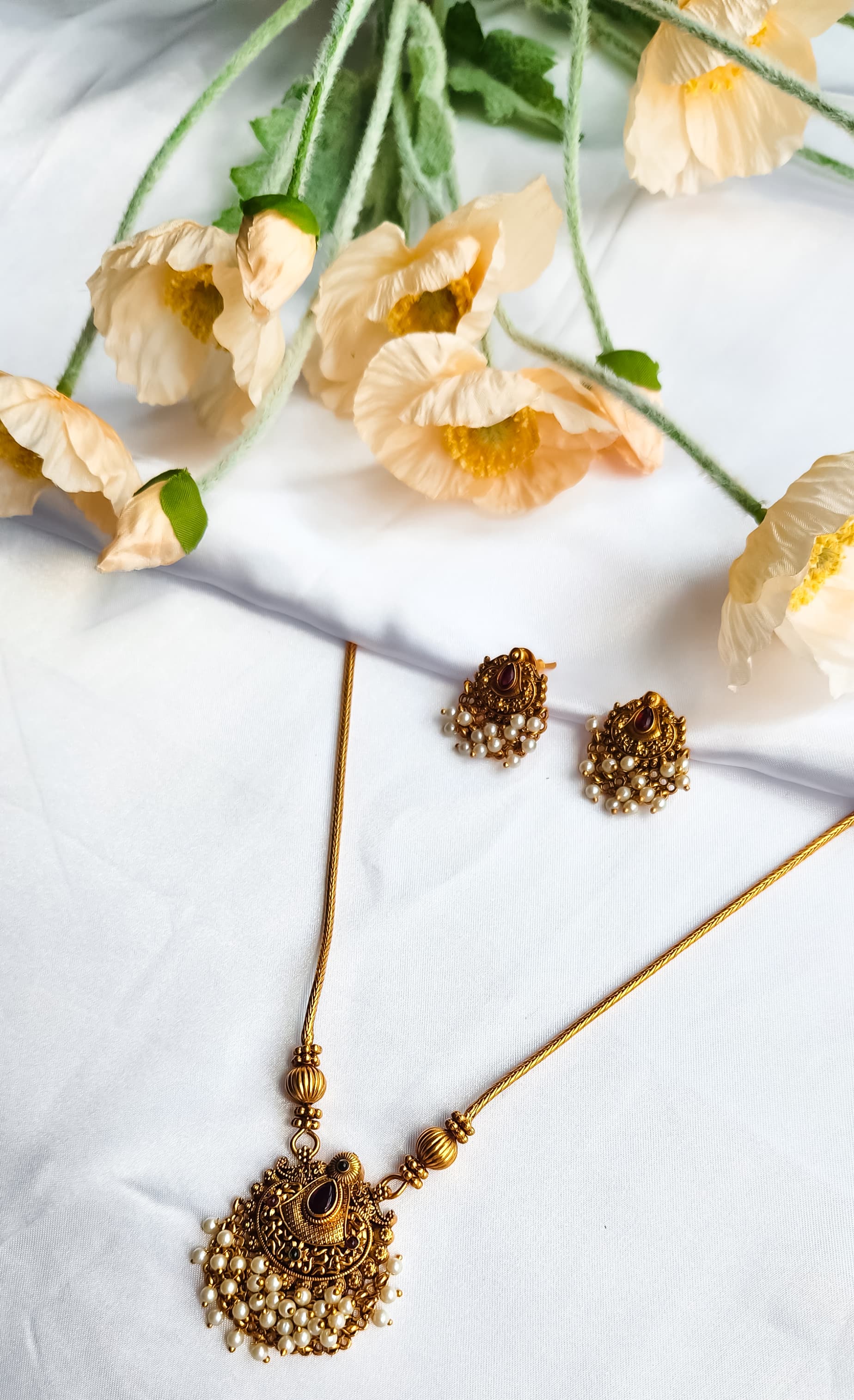 Antique gold temple-style necklace with pearl-detailed pendant and matching earrings on a white background with flowers.