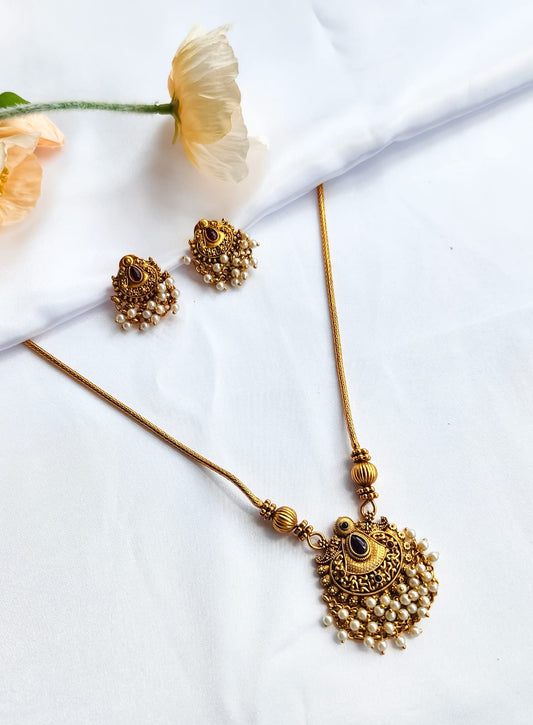 Antique gold temple-style necklace with pearl-detailed pendant and matching earrings on a white background with flowers.