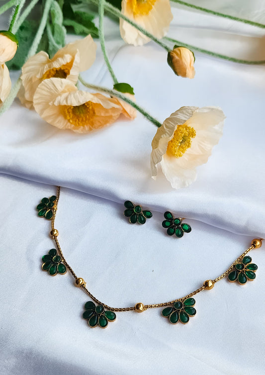 Green floral necklace with matching stud earrings displayed on white fabric with soft flowers in the background.